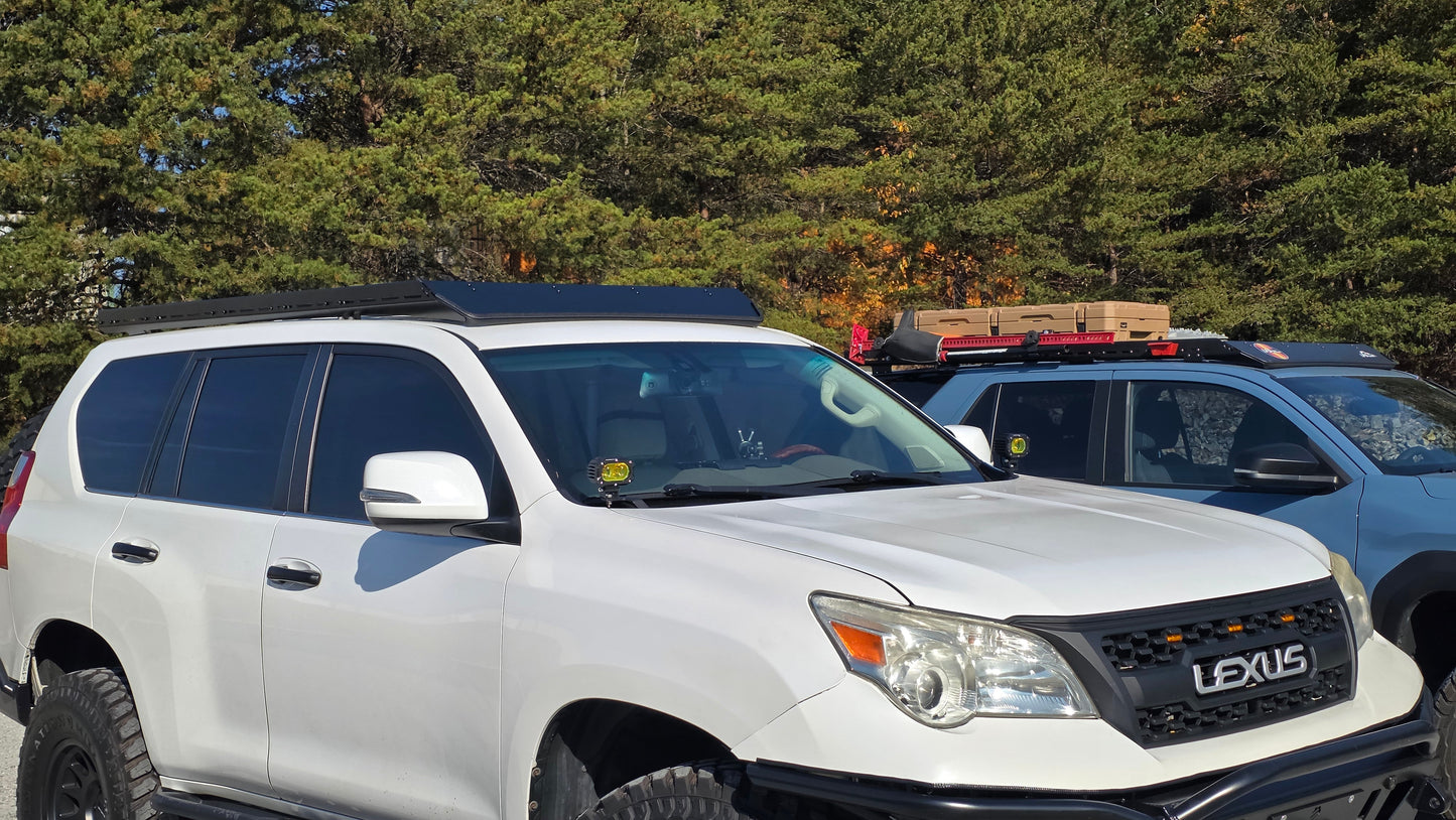 A white Lexus GX460 with a Ruggedized Low Profile Rack is parked beside a blue SUV with Heavy-Duty Crossbars and gear on its roof, both outdoors with green trees in the background.