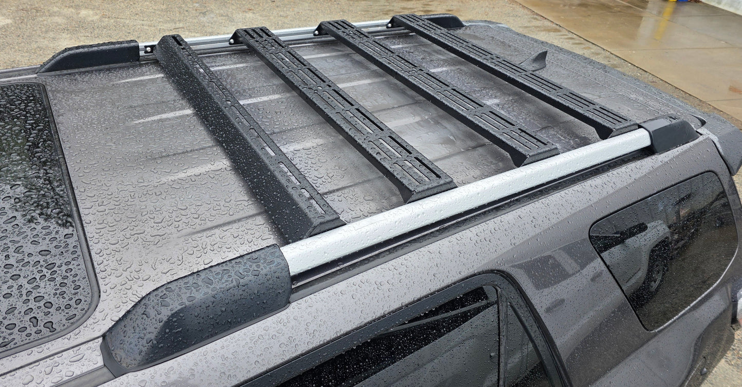 A close-up of a wet 4Runner roof shows the Wide Ruggedized Crossbar Kit - 5th Gen 4Runner covered in water droplets, with damp pavement below after rain. The black crossbars highlight the sturdy roof rack system.