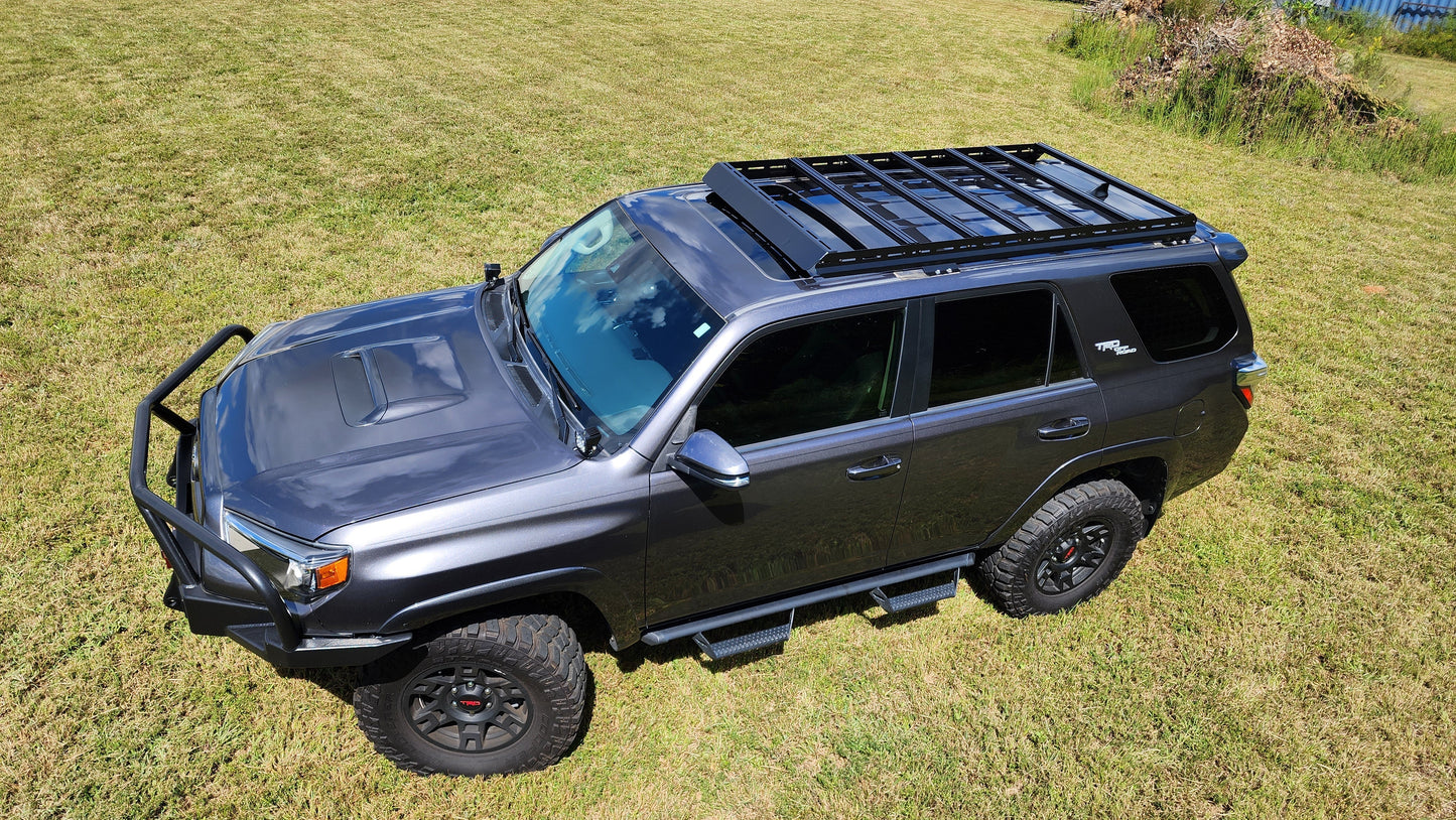 A matte black 5th Gen Toyota 4Runner with off-road tires, ruggedized crossbars, side steps, and the 7/8 Roof Rack - 5th Gen 4Runner is parked on green grass in a sunny outdoor area, ready for your next adventure.