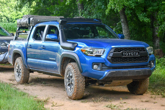 A blue Toyota Tacoma with off-road tires, a snorkel, and camping gear mounted on a Low Profile Extrusion Rack - 2nd and 3rd Gen Tacoma is parked on a dirt path surrounded by trees and greenery.