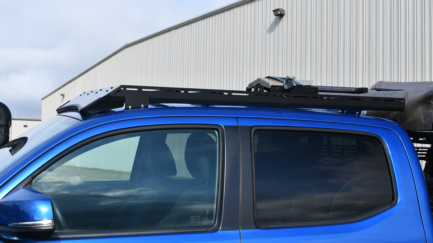 A blue 2nd or 3rd Gen Tacoma with a Low Profile Extrusion Rack and black crossbars is parked outside a large industrial building with metal siding, showing the side view of its front and rear doors and the mounted roof rack.