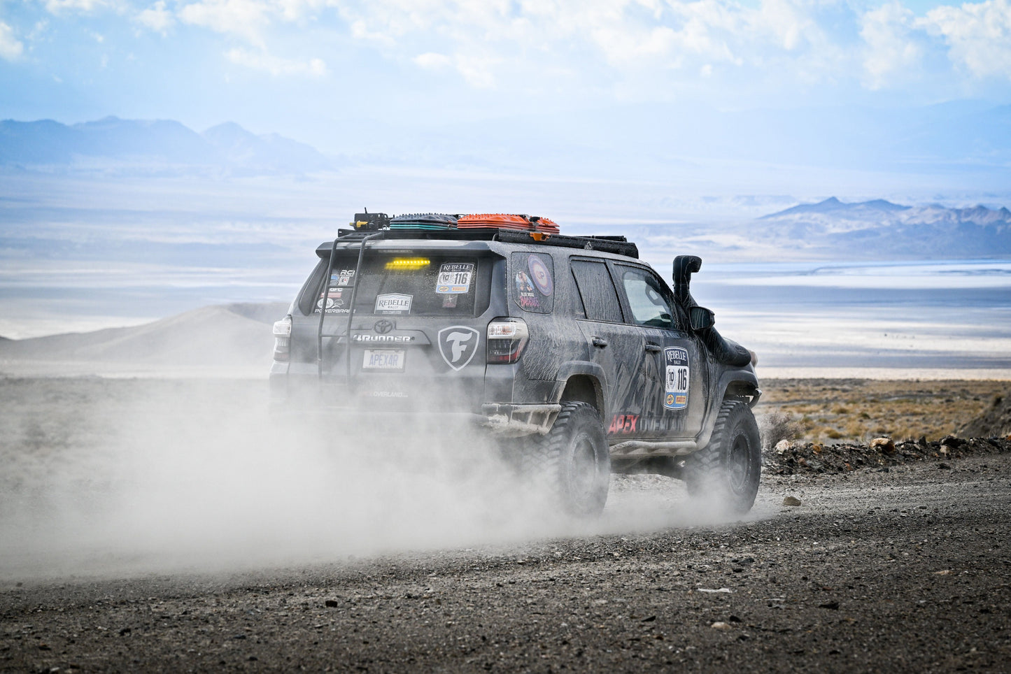 A 5th Gen 4Runner, fitted with the Ruggedized Low Profile Full Rack, kicks up dust on a remote mountain gravel road, its roof rack loaded and exterior marked by adventure under cloudy skies.