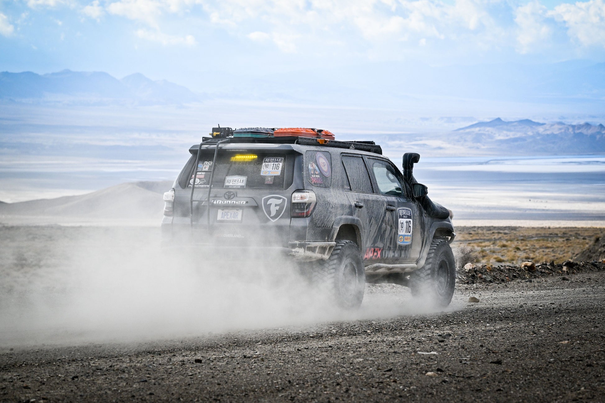 A 5th Gen 4Runner, fitted with the Ruggedized Low Profile Full Rack, kicks up dust on a remote mountain gravel road, its roof rack loaded and exterior marked by adventure under cloudy skies.