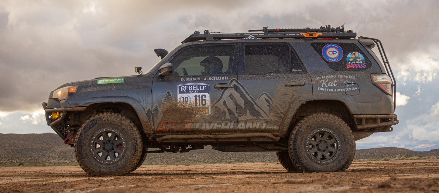 A 5th Gen 4Runner featuring a Ruggedized Low Profile Full Rack, large tires, heavy-duty crossbars, and mountain-themed decals with the number 116 is parked on dirt with mountains and a cloudy sky in the background.