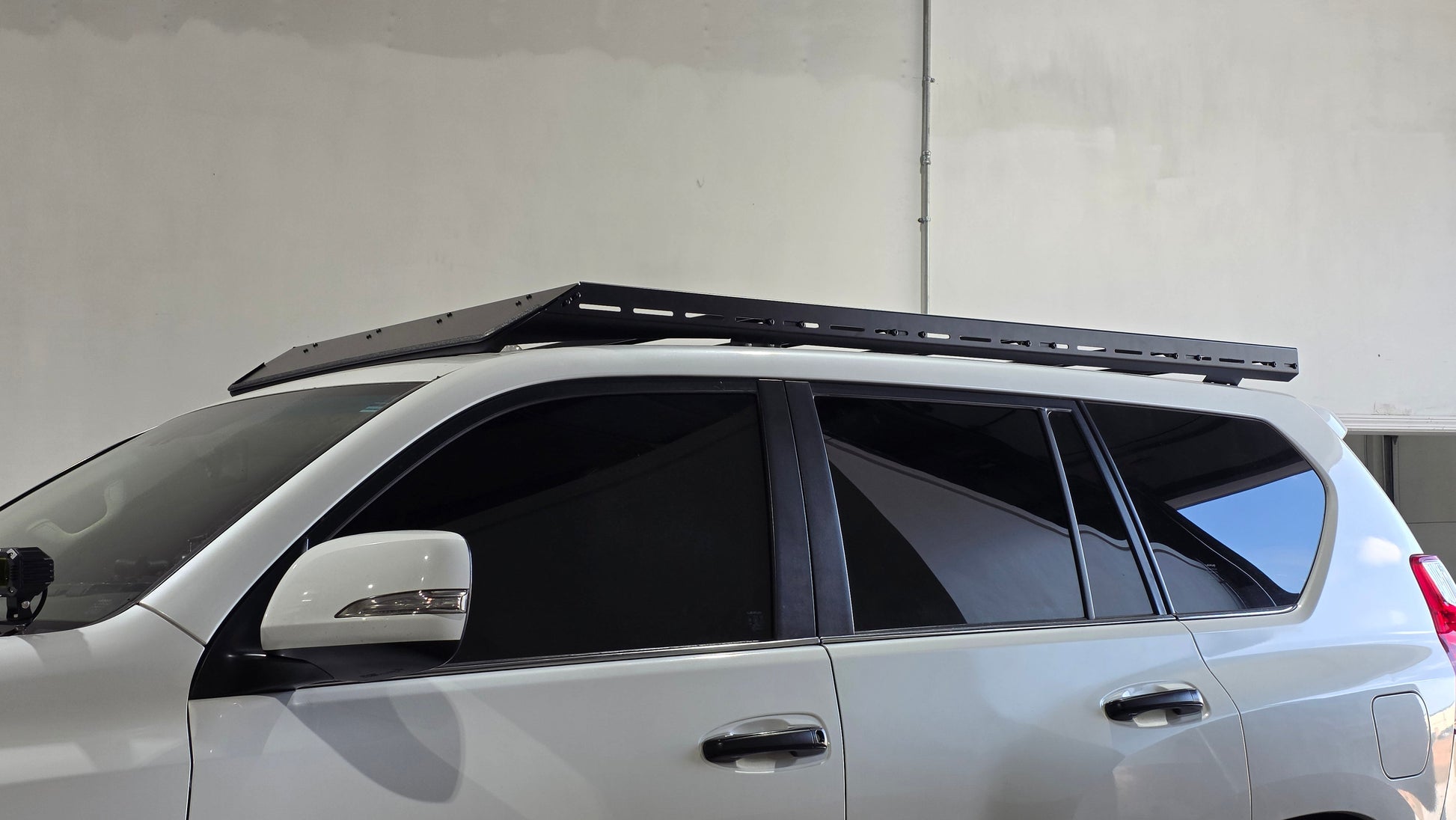 A white SUV with tinted windows is parked indoors, featuring a Ruggedized Low Profile Rack - GX460 with heavy-duty crossbars mounted on the roof. A plain light-colored wall is visible in the background.