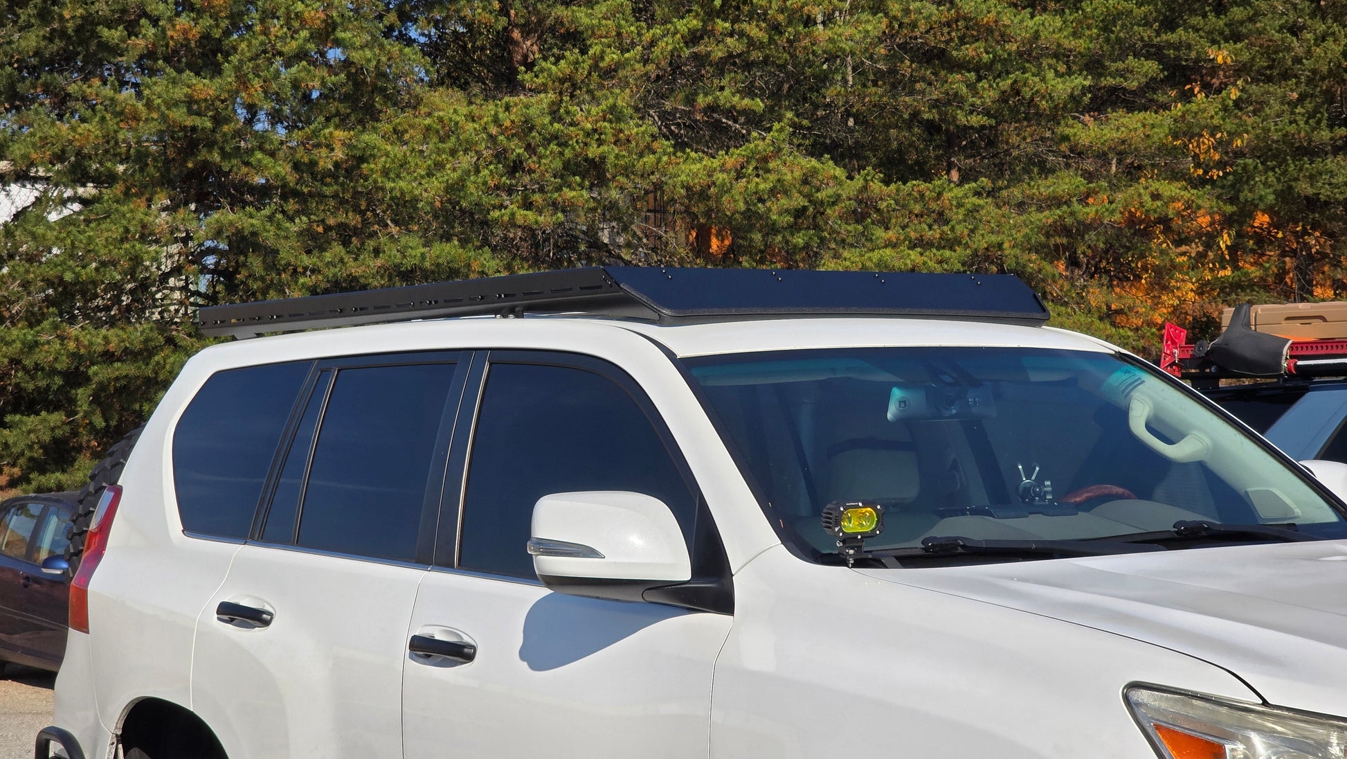 A white SUV with dark tinted windows and a Ruggedized Low Profile Rack - GX460 featuring heavy-duty crossbars is parked outdoors near trees. Sunlight reflects off the car, and a small yellow ornament sits on the dashboard.