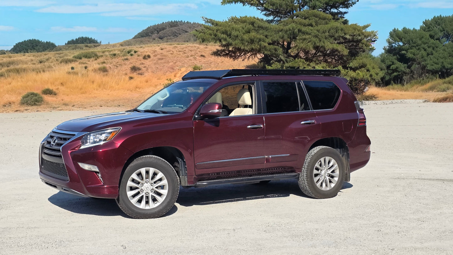A maroon Lexus GX460 with a Ruggedized Low Profile Rack is parked on gravel, surrounded by dry grass, green trees, and hills beneath a partly cloudy blue sky.