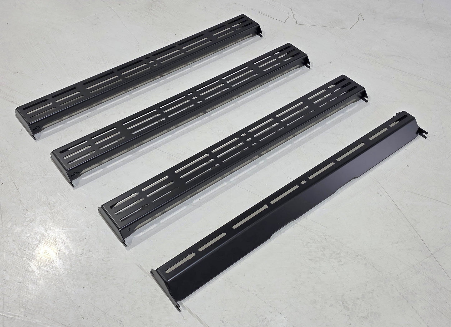 Four wide, black metal panels from the Wide Ruggedized Crossbar Kit - 5th Gen 4Runner are aligned parallel on a light gray floor. Three feature multiple slots, while one is solid with a few cutouts.