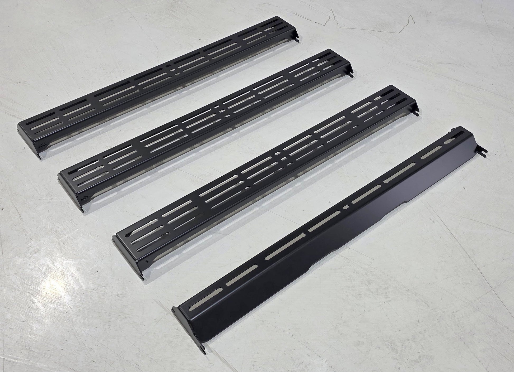 Four wide, black metal panels from the Wide Ruggedized Crossbar Kit - 5th Gen 4Runner are aligned parallel on a light gray floor. Three feature multiple slots, while one is solid with a few cutouts.
