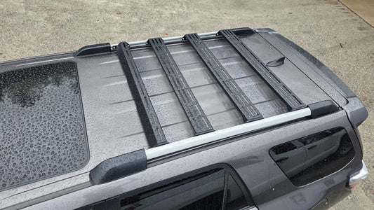 Top view of a gray 5th Gen 4Runner with rain droplets on the roof and a Wide Ruggedized Crossbar Kit - 5th Gen 4Runner mounted to the roof rails, parked on wet concrete.