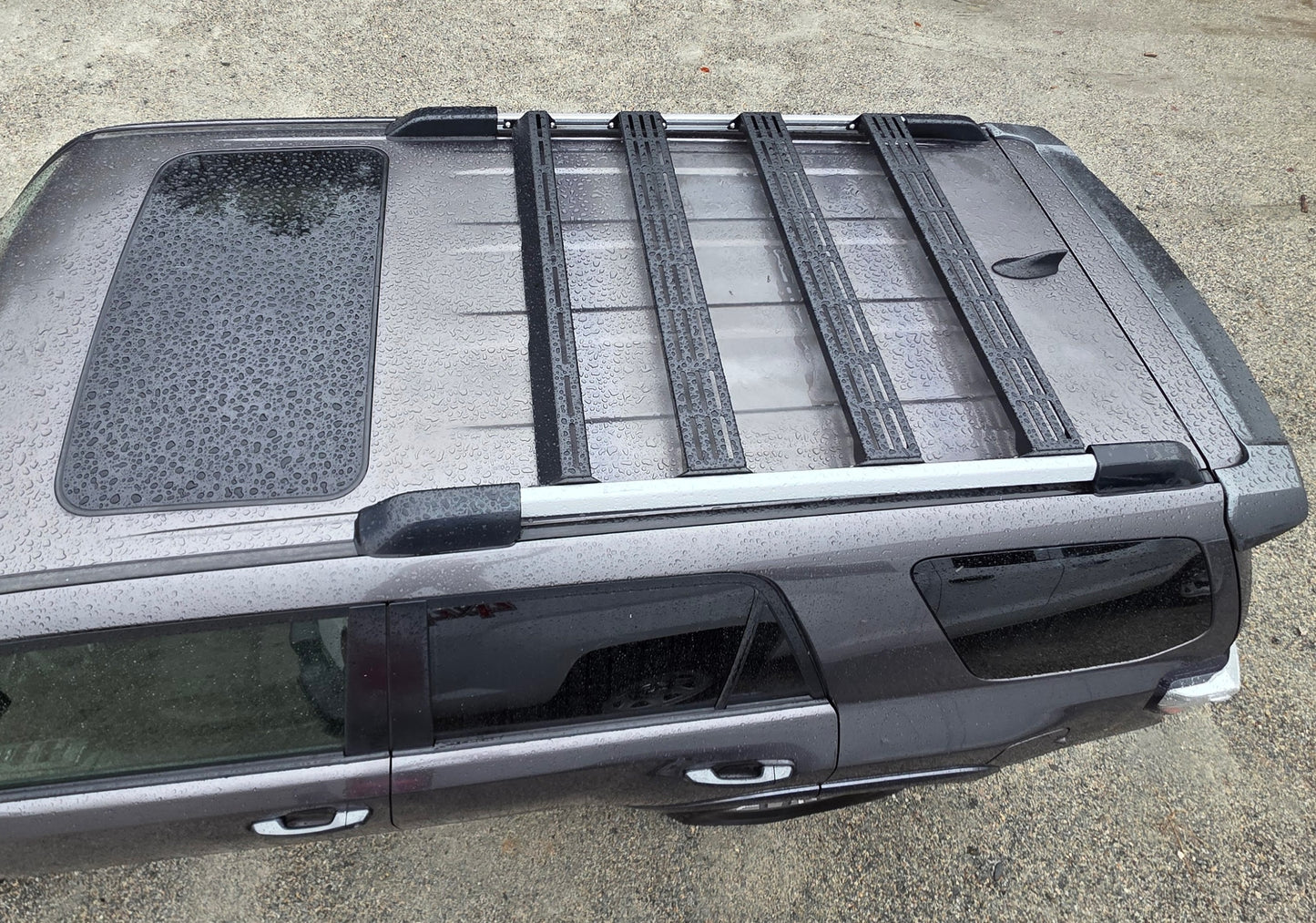 A top-down view of a gray 5th Gen 4Runner with a sunroof and the Wide Ruggedized Crossbar Kit installed. The SUV is parked on gravel, and raindrops are visible on the roof.