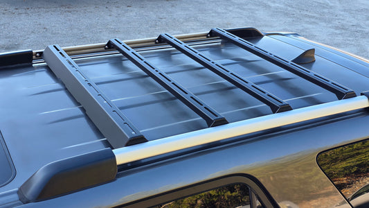 Close-up of a 5th Gen 4Runner with the Ruggedized Crossbar Kit installed—showing four black crossbars mounted on factory roof rails against a blurred outdoor background, offering an ideal roof storage solution.