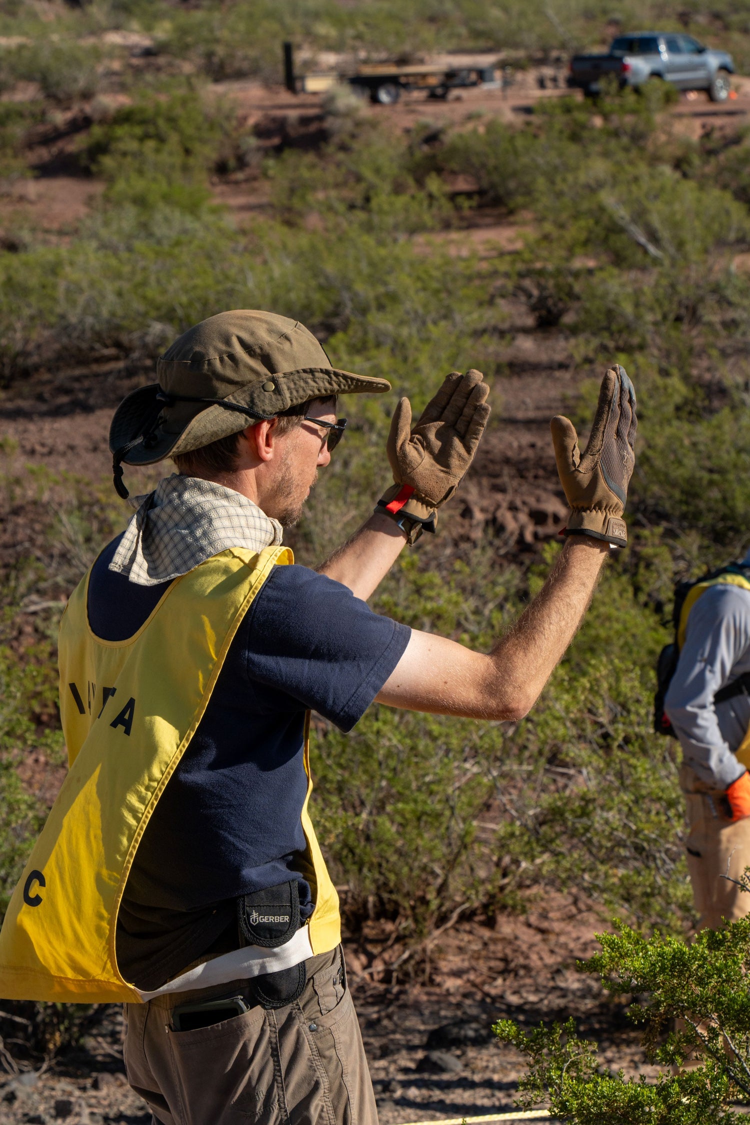 A person wearing a yellow vest, gloves, and a hat gestures with both hands outdoors in a desert-like area, while another person in the background stands partially out of frame. Shrubs and a vehicle are visible in the distance.