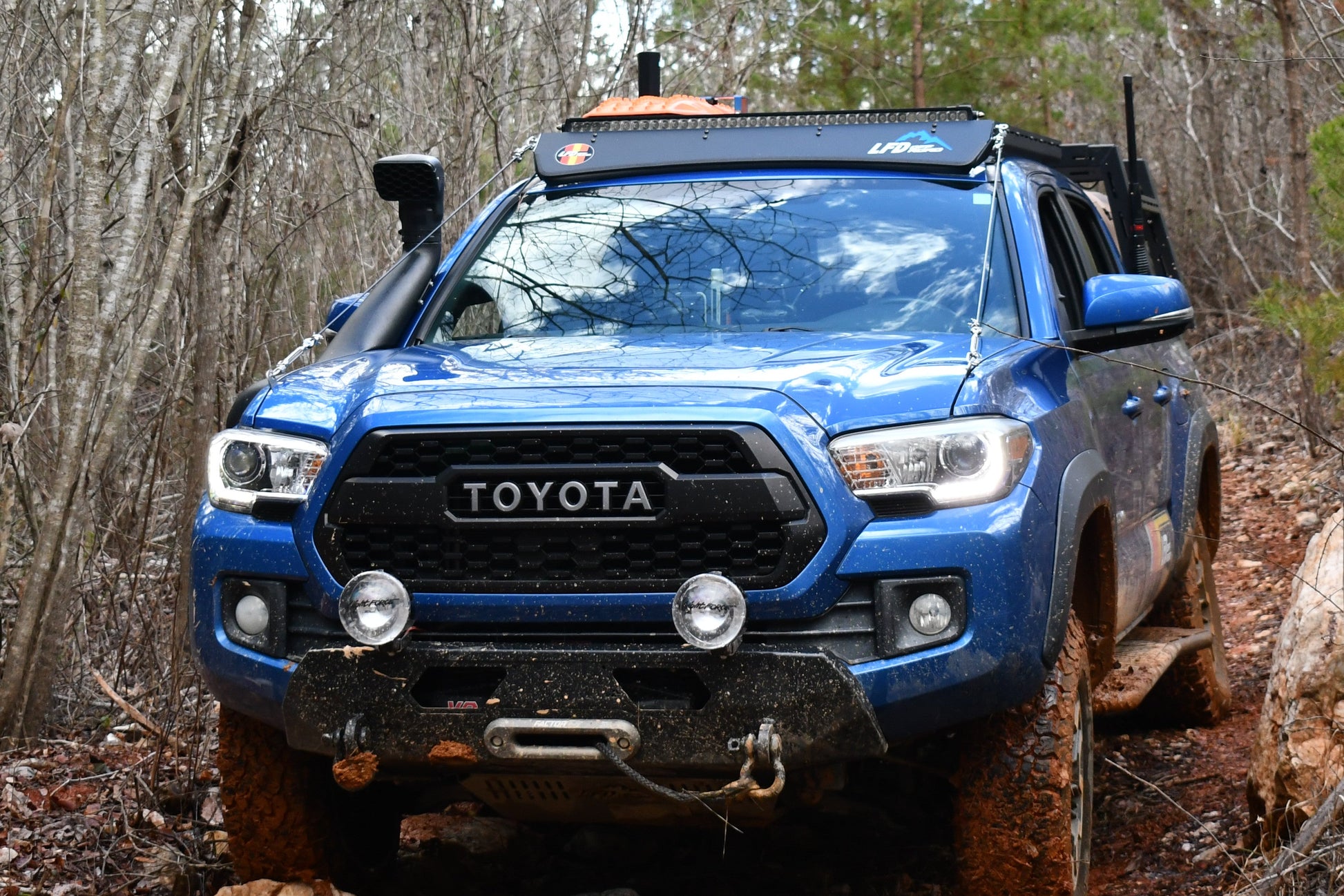 A blue Toyota Tacoma with 3rd Gen Tacoma Limb Riser Hood Brackets and off-road gear tackles a muddy, rocky forest trail surrounded by leafless trees and evergreens.