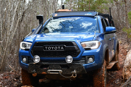 A blue Toyota Tacoma with 3rd Gen Tacoma Limb Riser Hood Brackets and off-road gear tackles a muddy, rocky forest trail surrounded by leafless trees and evergreens.