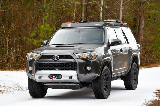 A gray Toyota 4Runner with black wheels and a Ruggedized Low Profile Full Rack - 5th Gen 4Runner is parked on a snowy road among trees, featuring off-road upgrades like a winch-equipped front bumper and red tow hook.
