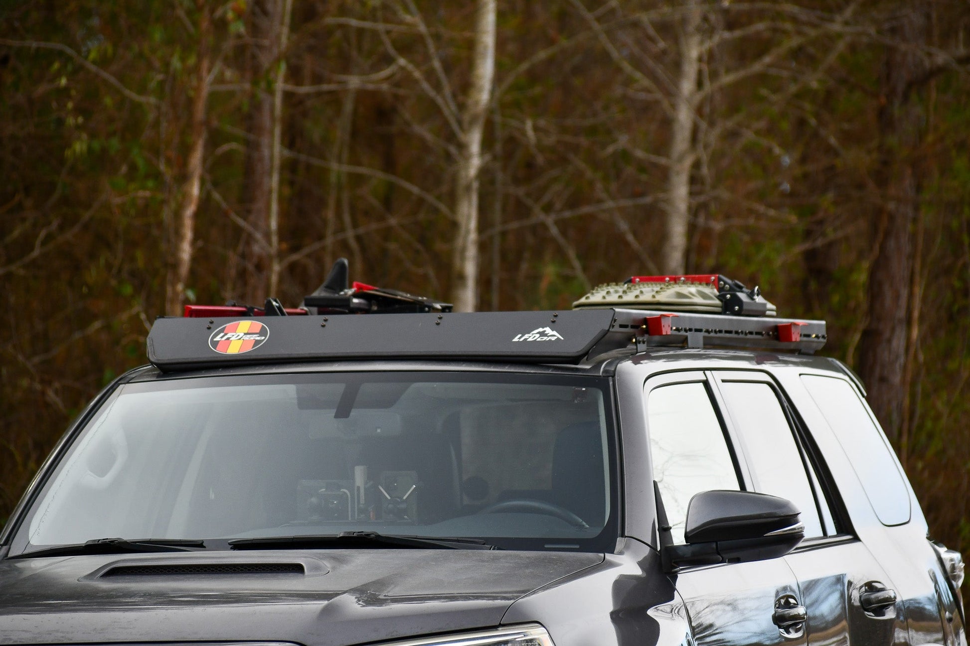 A close-up of a 5th Gen 4Runner fitted with the Ruggedized Low Profile Full Rack, carrying gear like a shovel and storage container, parked in front of leafless trees in a wooded area.