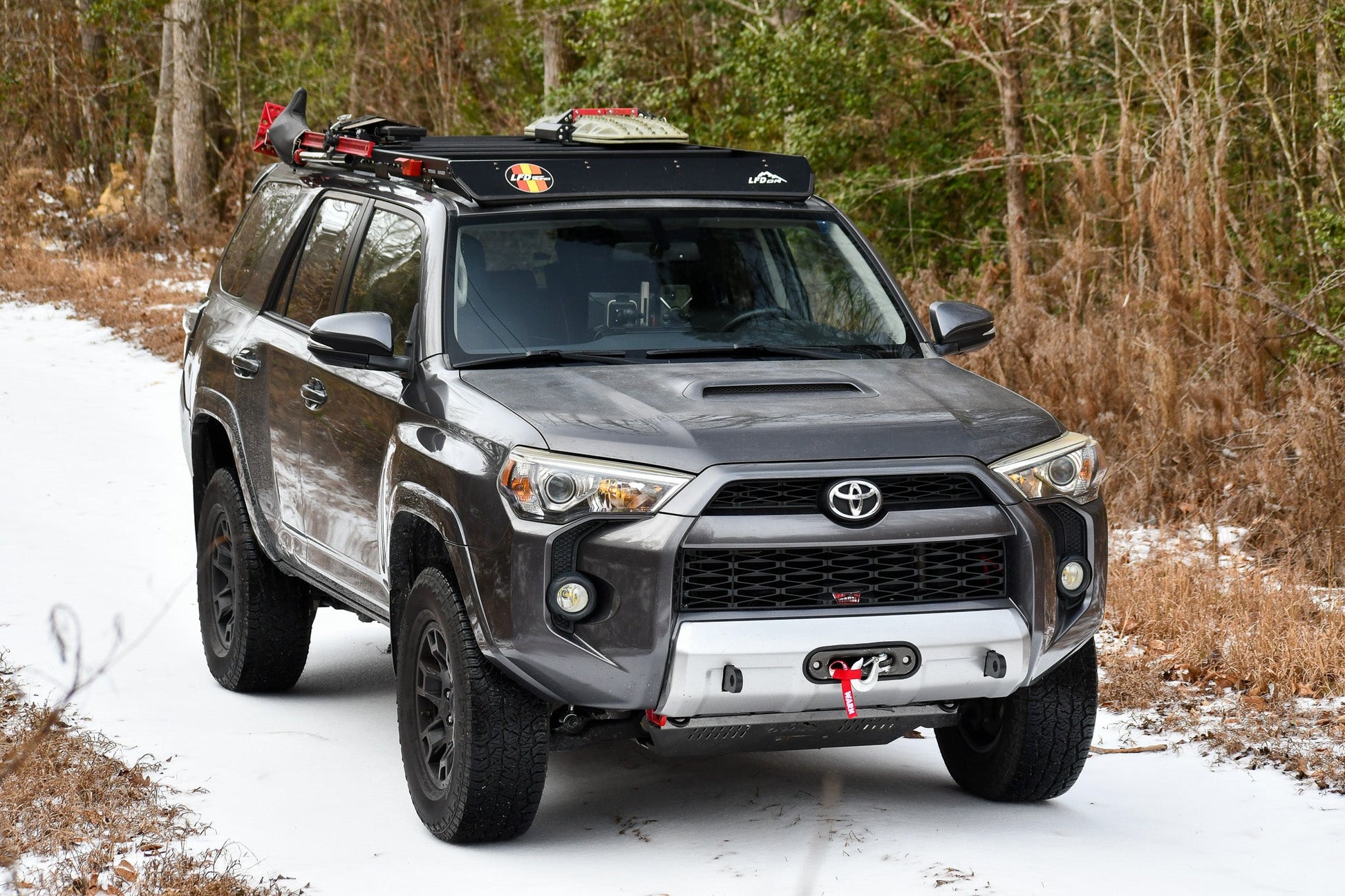 A gray 5th Gen Toyota 4Runner with a Ruggedized Low Profile Full Rack is parked on a snowy trail, sporting heavy-duty crossbars loaded with gear, black wheels, and a front skid plate among snowy patches and brown-leaved trees.
