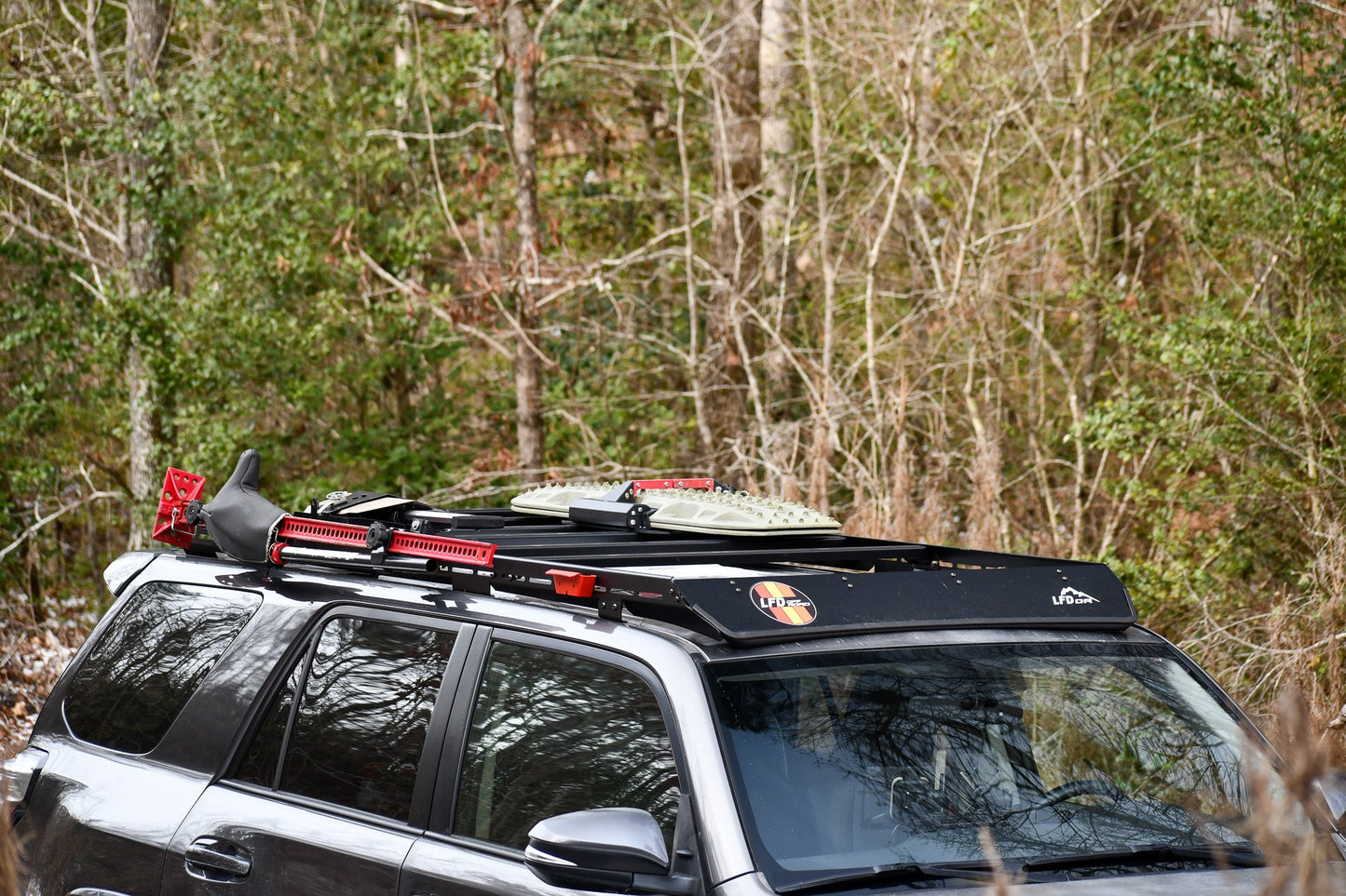 A dark 5th Gen 4Runner with a Ruggedized Low Profile Full Rack is parked by dense forest, carrying off-road gear like a shovel, traction boards, and a red jack securely mounted on the heavy-duty crossbars.