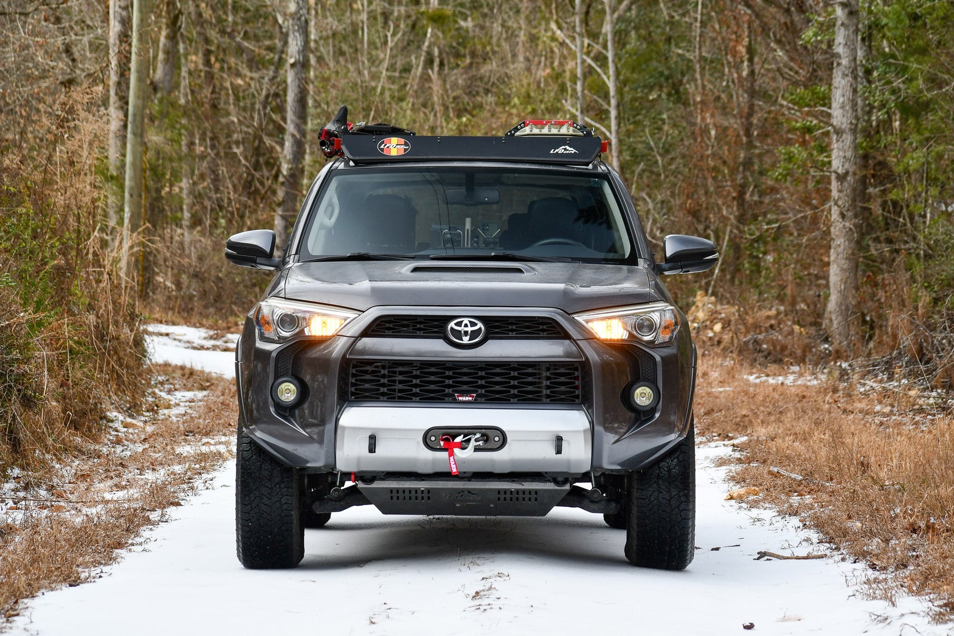 A gray 5th Gen Toyota 4Runner with a Ruggedized Low Profile Full Rack is parked on a narrow, snow-dusted dirt path among leafless trees and dry grass. The headlights are on, and there's a winch on the front bumper.