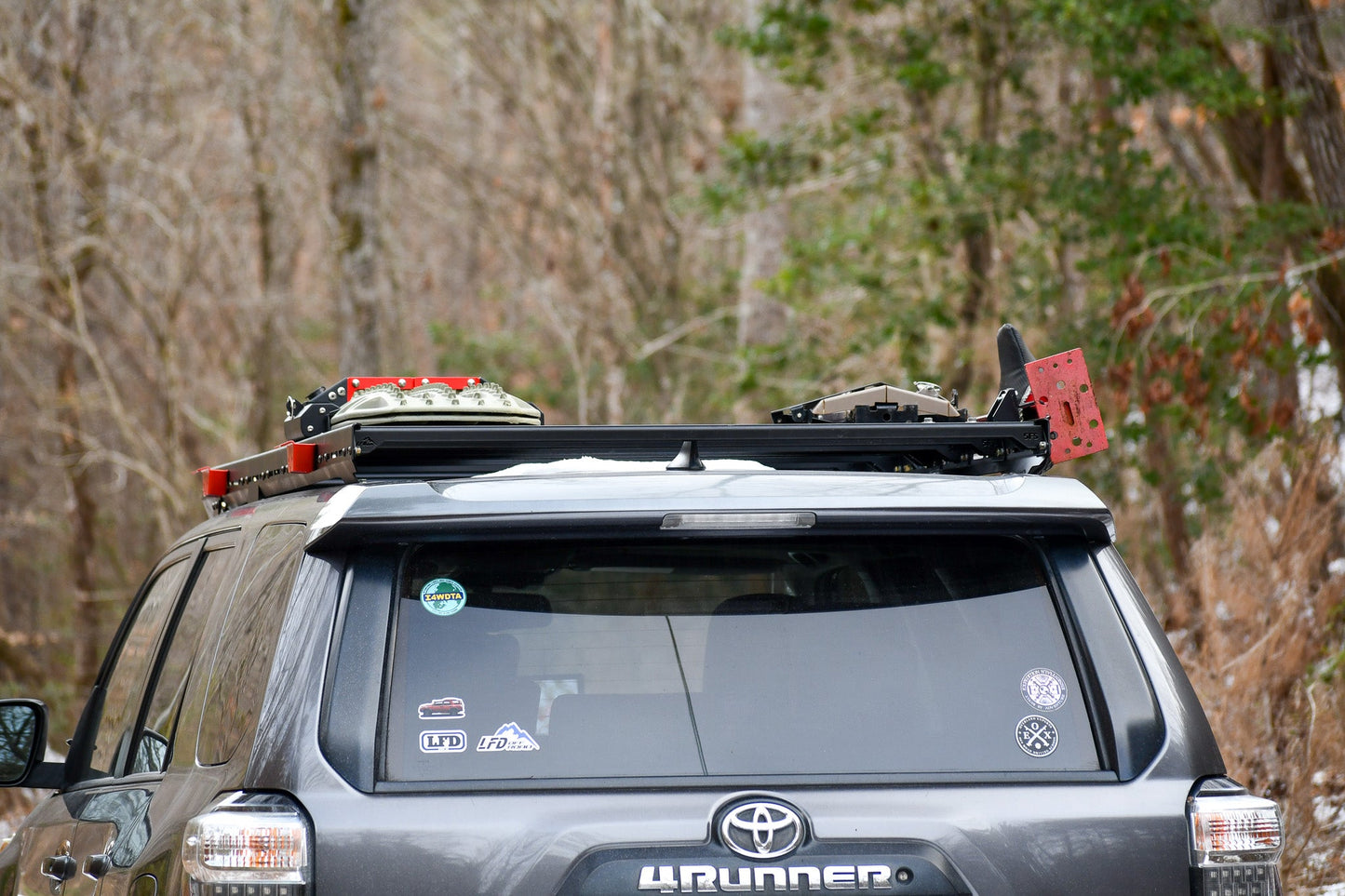 A gray Toyota 4Runner with a Ruggedized Low Profile Full Rack - 5th Gen 4Runner holds traction boards, a shovel, and a red fuel can on top. Stickers cover the rear window, and trees fill the outdoor background.