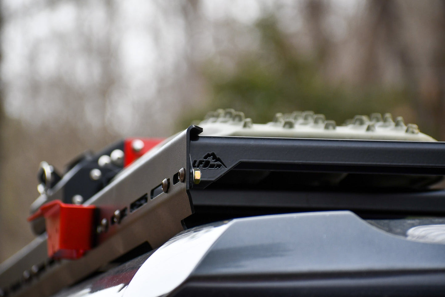 Close-up of off-road gear on a Ruggedized Low Profile Full Rack - 5th Gen 4Runner, featuring a red tool and green recovery track mounted to heavy-duty crossbars, against a blurred natural backdrop.