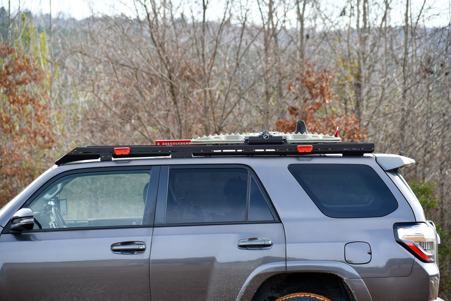 A grey 5th Gen 4Runner with a Ruggedized Low Profile Full Rack is parked outdoors, showing heavy-duty crossbars and recovery boards mounted on top. Leafless trees and a cloudy sky appear in the background.