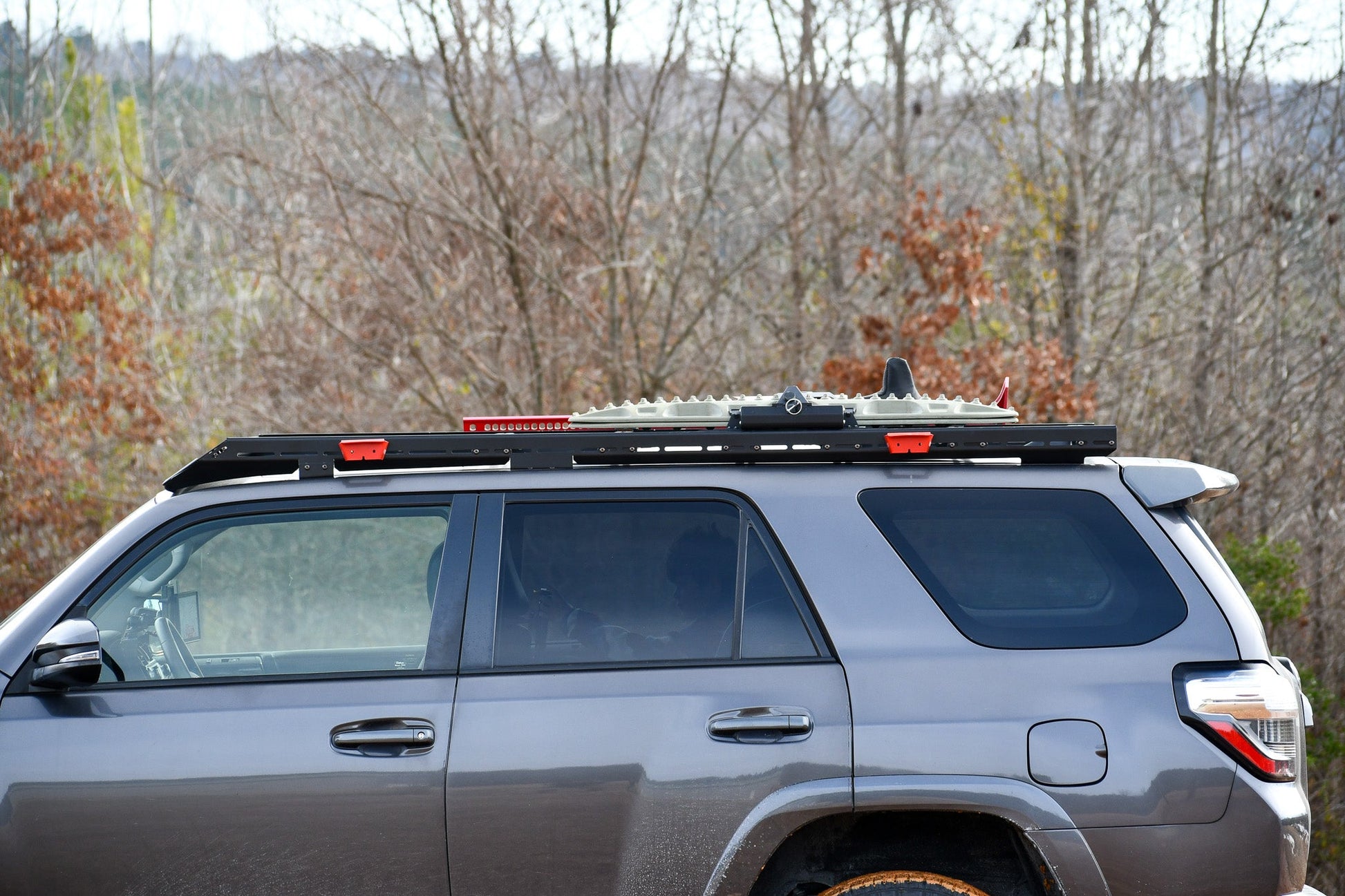 A grey 5th Gen 4Runner with a Ruggedized Low Profile Full Rack is parked outdoors, showing heavy-duty crossbars and recovery boards mounted on top. Leafless trees and a cloudy sky appear in the background.