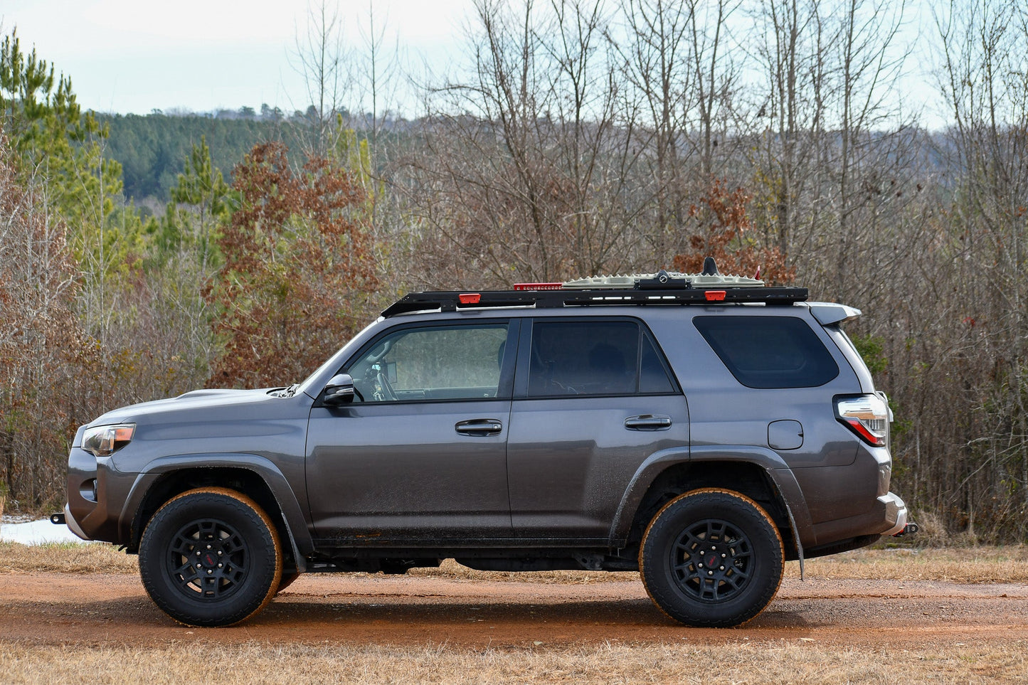 A 5th Gen 4Runner equipped with the Ruggedized Low Profile Full Rack is parked on a dirt road among leafless trees and evergreens, with hills in the background.