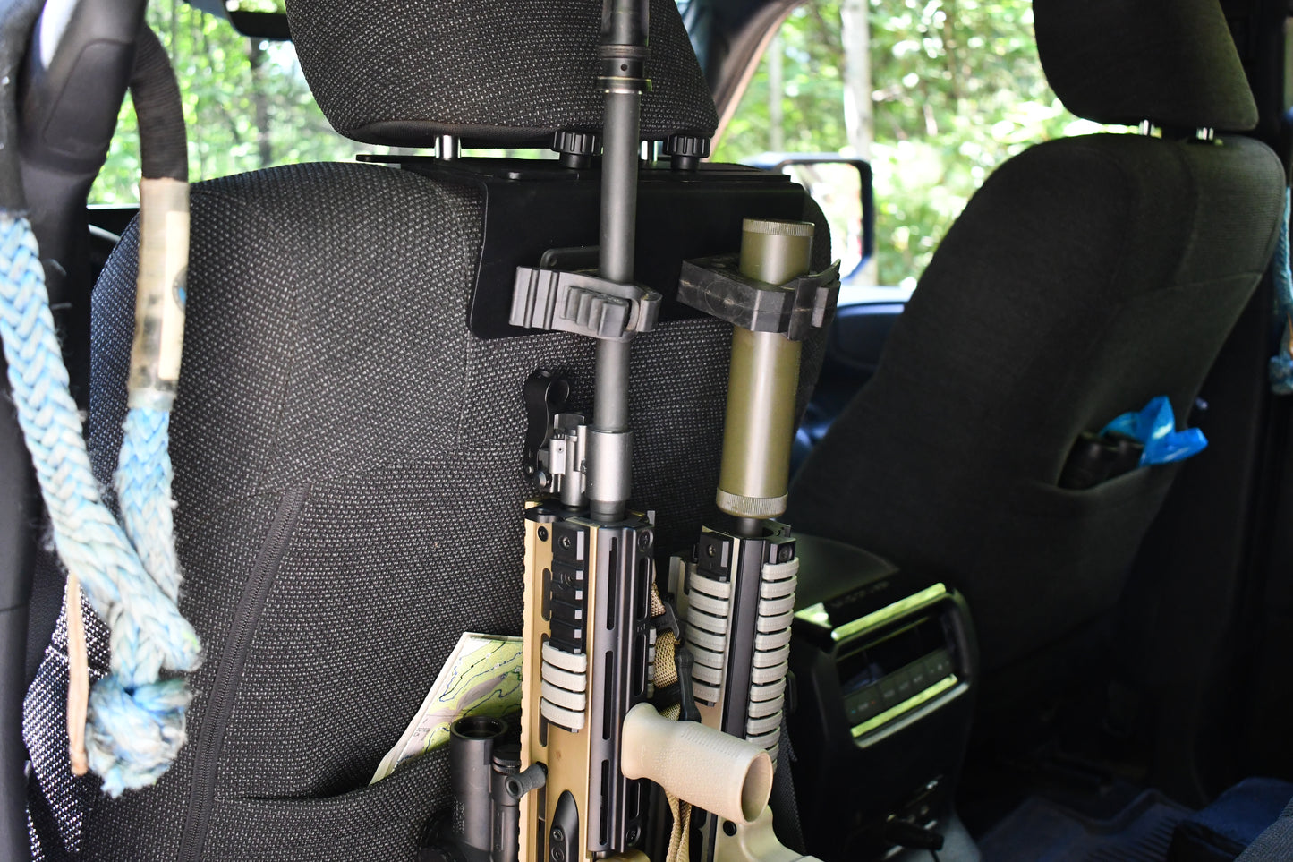 Two rifles with tactical attachments are secured behind the front passenger seat of a vehicle using the Headrest Rifle Mount - HRM, with a rope visible on the left and green foliage outside the window.
