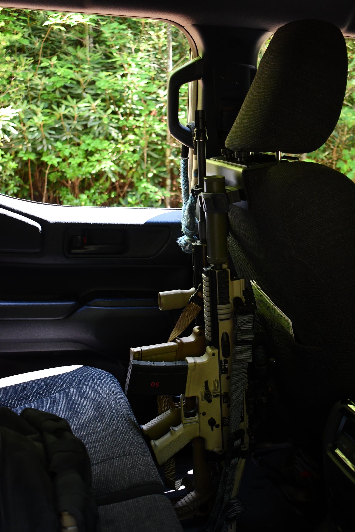 A tan rifle is secured behind the passenger seat with a Headrest Rifle Mount - HRM inside a vehicle, while lush green foliage can be seen outside the window.