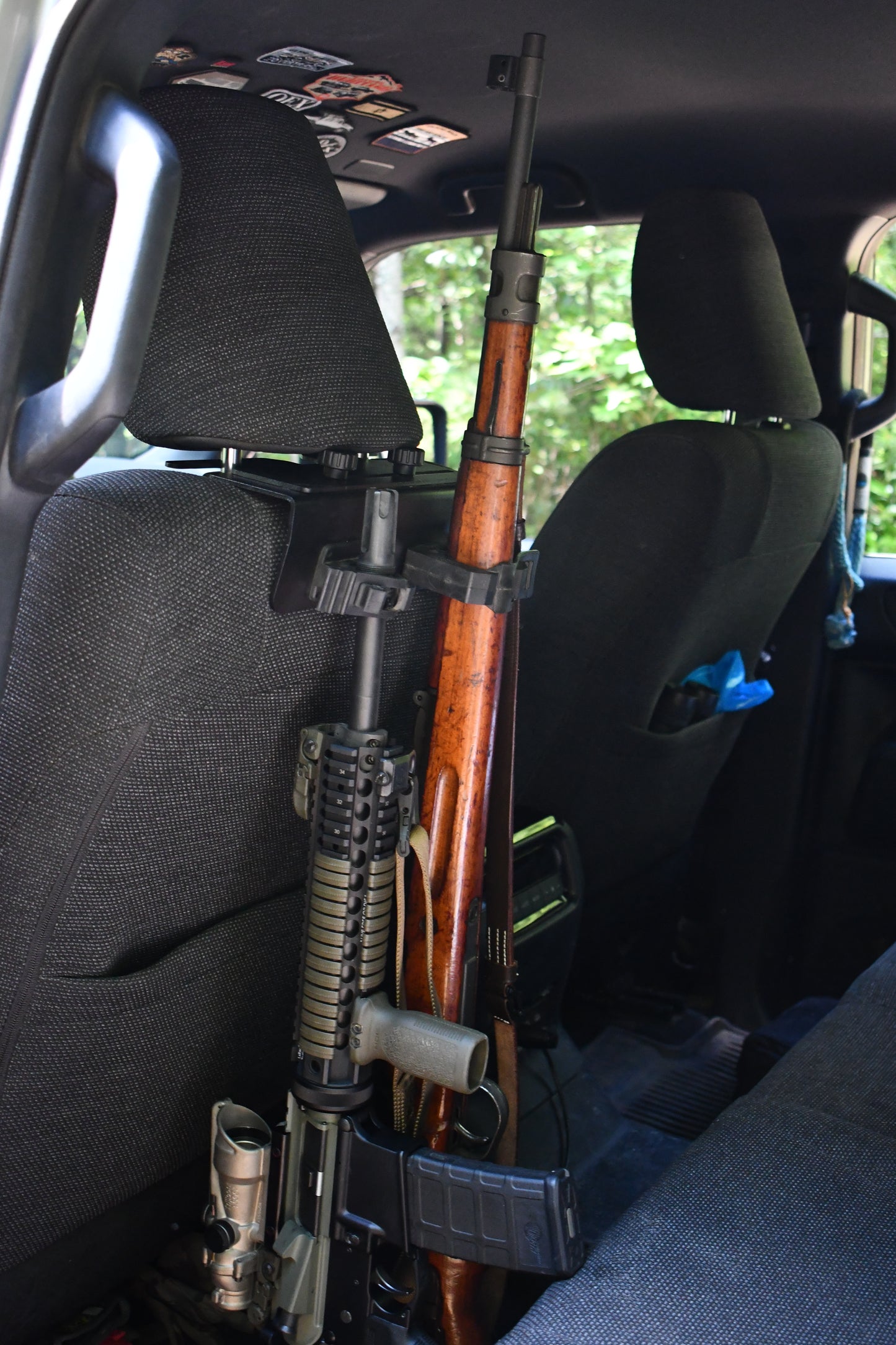 Two rifles, one with a wooden stock and one tactical, are secured in a Headrest Rifle Mount - HRM attached to the back of a front car seat. Sunlight streams through the window, revealing greenery outside.
