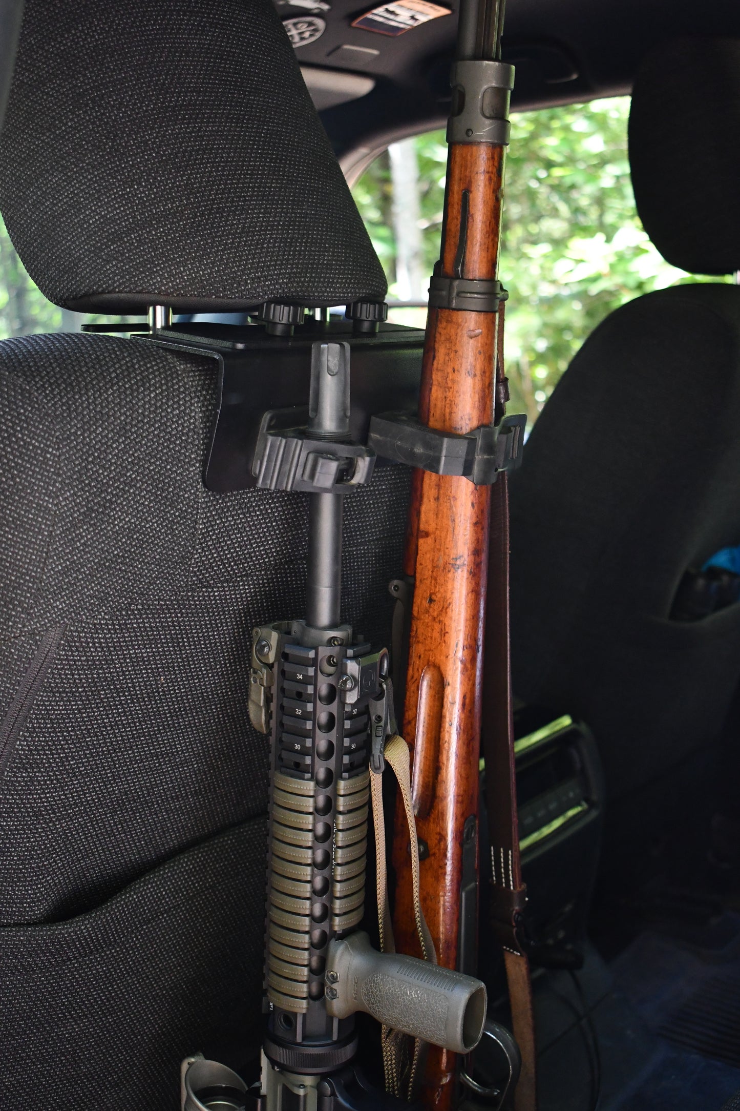 A close-up of two rifles secured by the Headrest Rifle Mount - HRM on a car seat back, with forest greenery visible through the window.