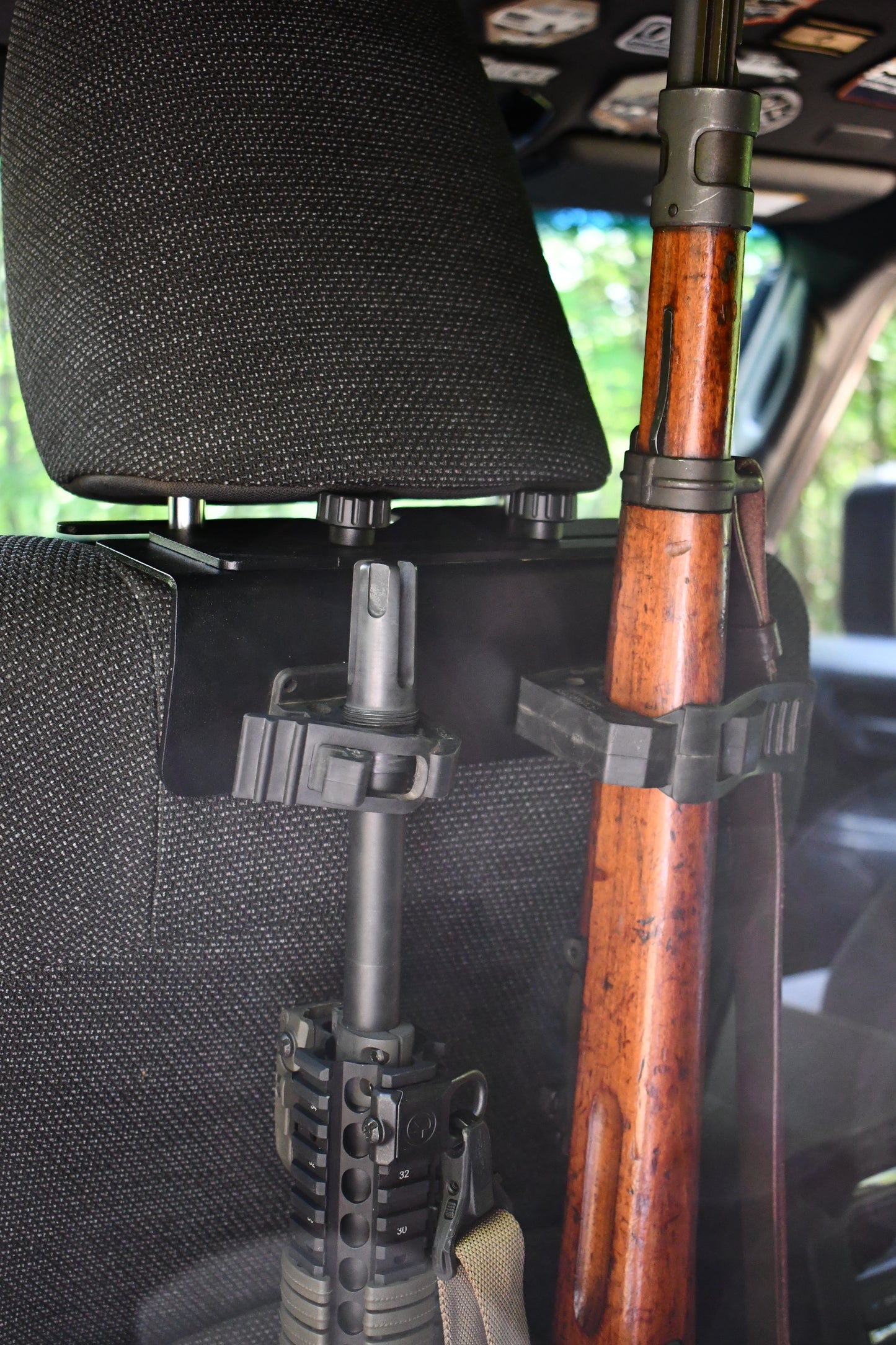 Two rifles are secured in a gun rack using the Headrest Rifle Mount - HRM, attached to the back of a vehicle seat with the headrest and part of the vehicle interior visible.