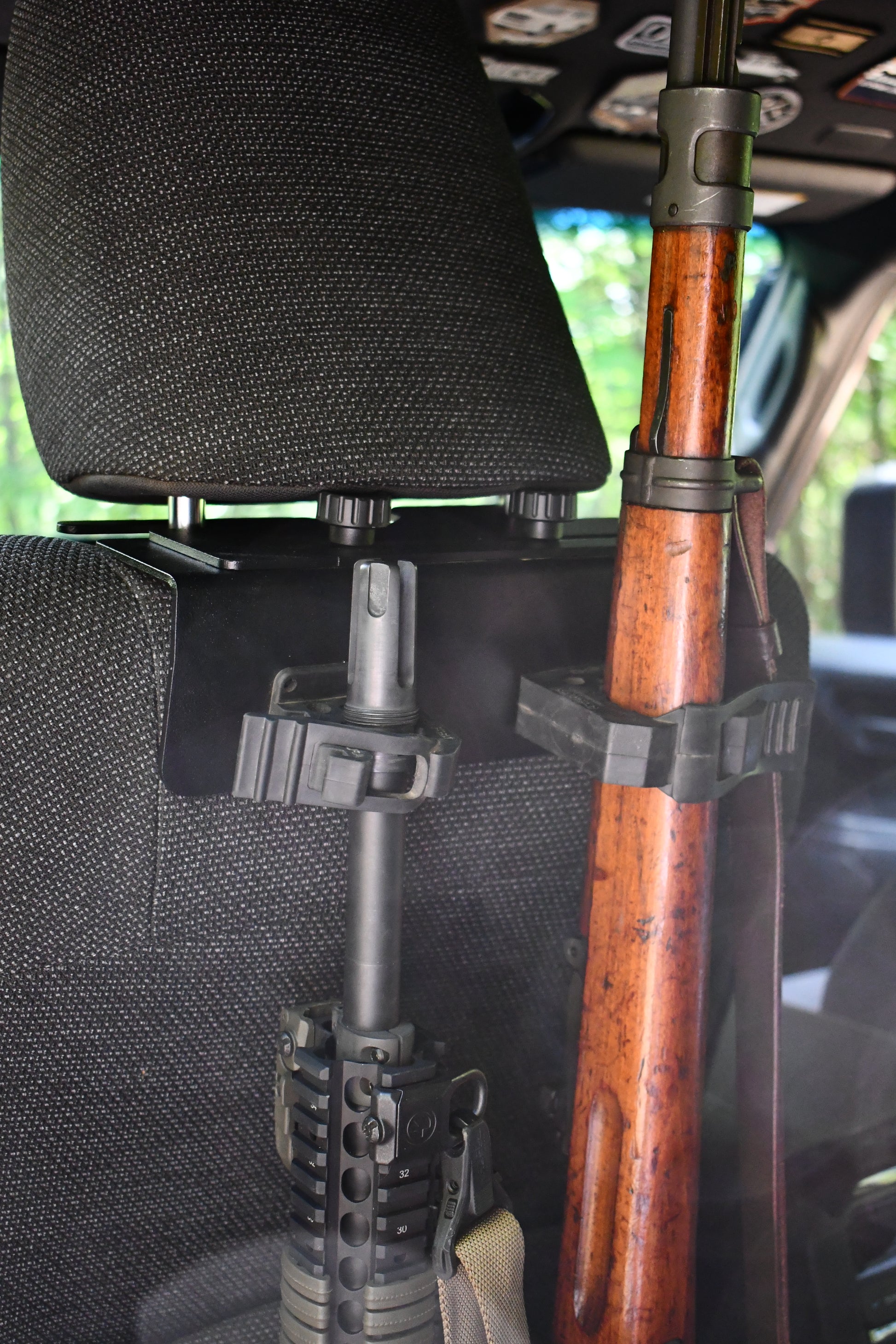 Two rifles are secured in a gun rack using the Headrest Rifle Mount - HRM, attached to the back of a vehicle seat with the headrest and part of the vehicle interior visible.