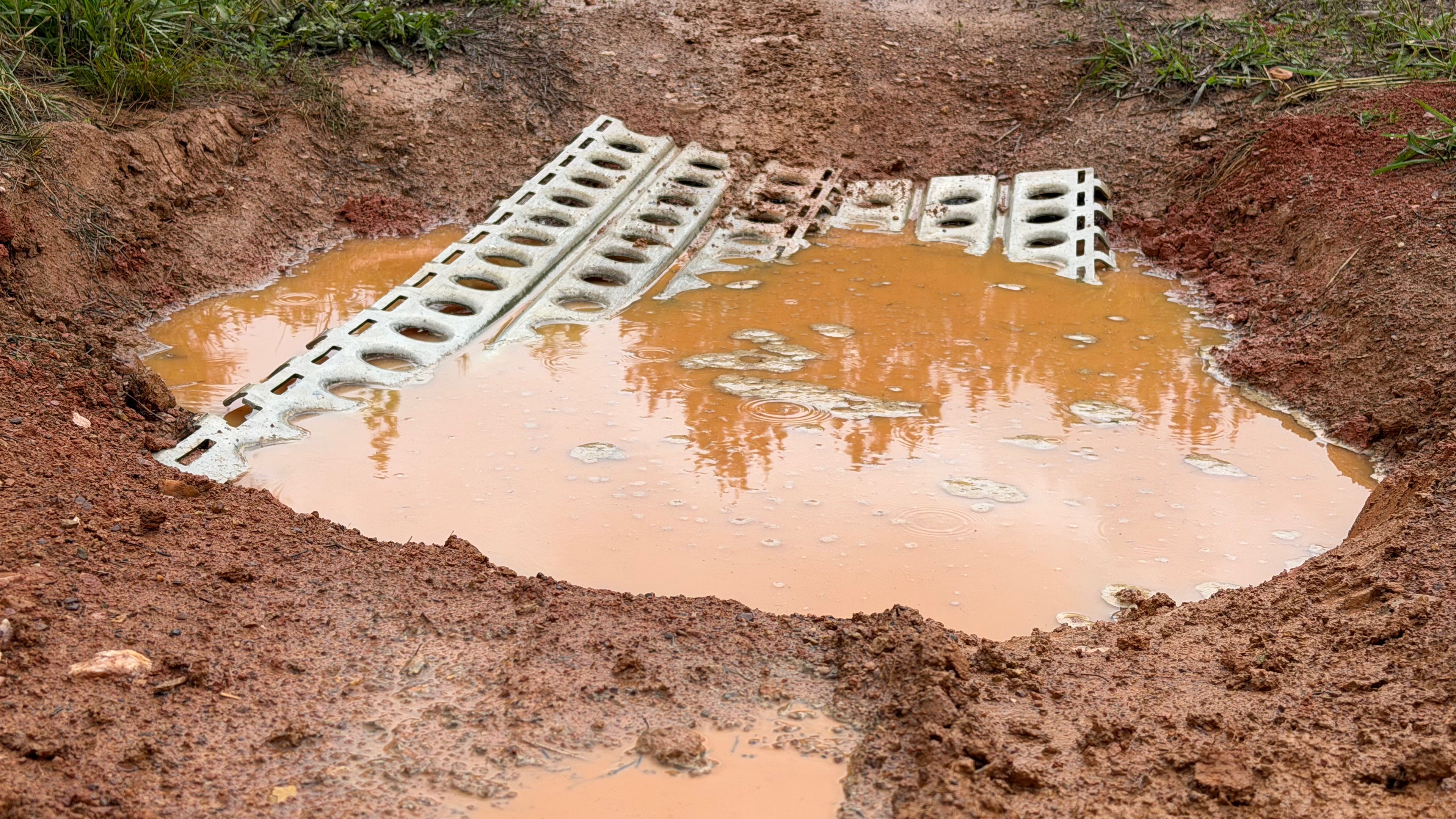 A muddy, water-filled rut in a dirt road with two white traction boards laid across the puddle to help vehicles pass through. Grass and wet soil surround the area.