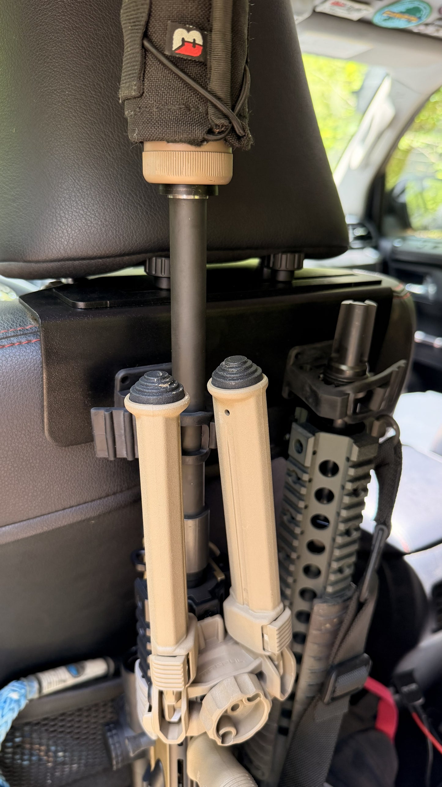 A close-up of a car seat back with tactical gear, including a tan bipod, part of a firearm secured in the Headrest Rifle Mount - HRM, and a patch case inside the sunlit vehicle interior.