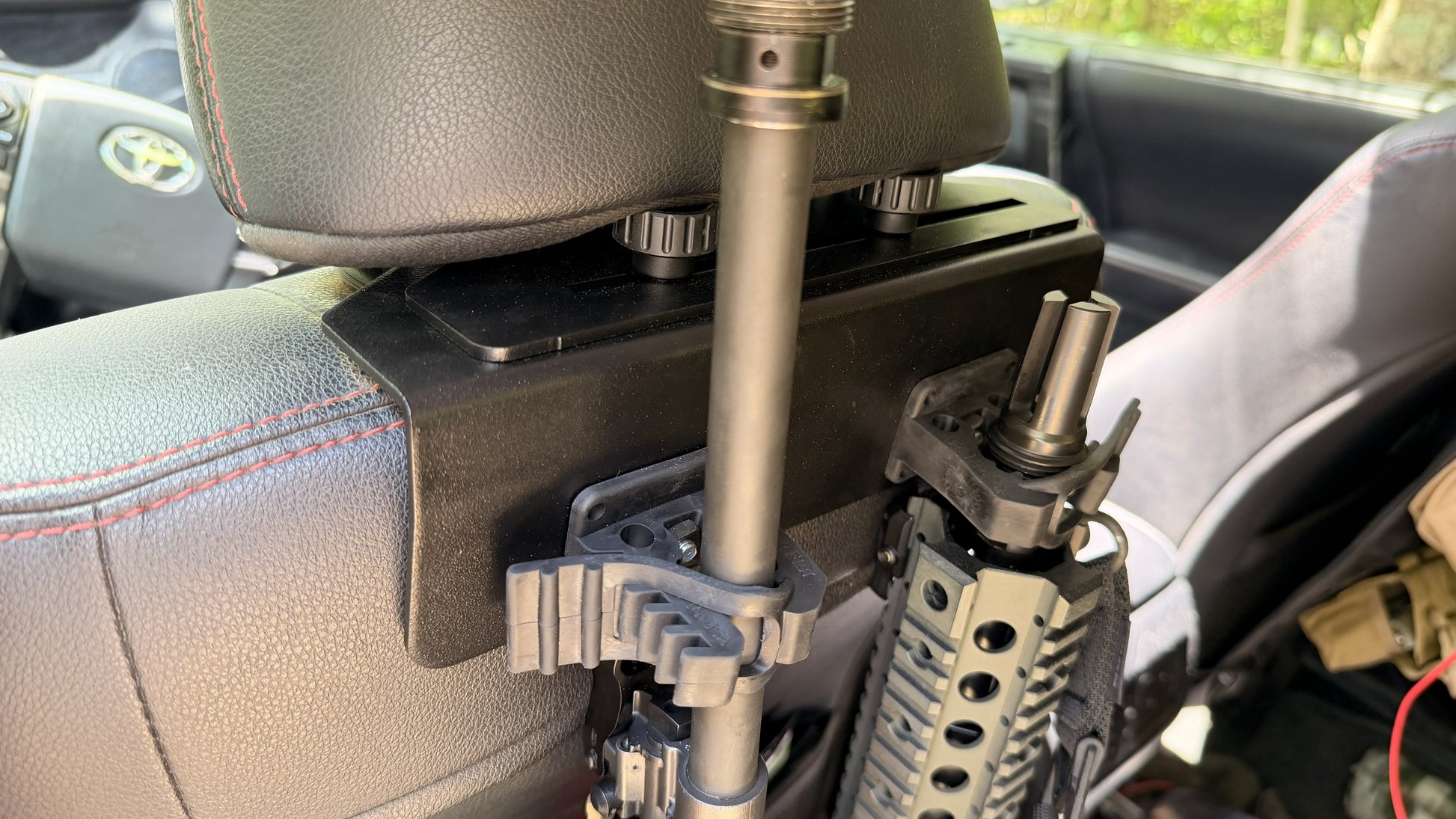 Two rifles secured in a Headrest Rifle Mount - HRM attached to the back of a car seat, with part of the vehicle's interior visible.