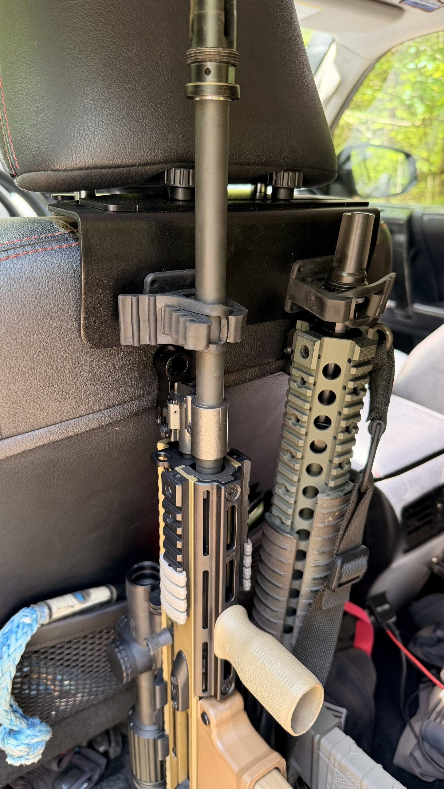 Two rifles are secured vertically behind a car seat using the Headrest Rifle Mount - HRM. The photo, taken inside a vehicle, shows the rifles' barrels and handguards positioned near the seatback for safe transport.
