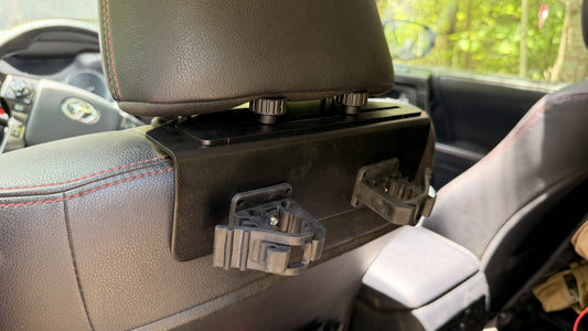 A close-up of the Headrest Rifle Mount - HRM, fitted to a car’s front seat headrest with two rifle quickfist clamps, positioned above the back seat, forest visible through the window.
