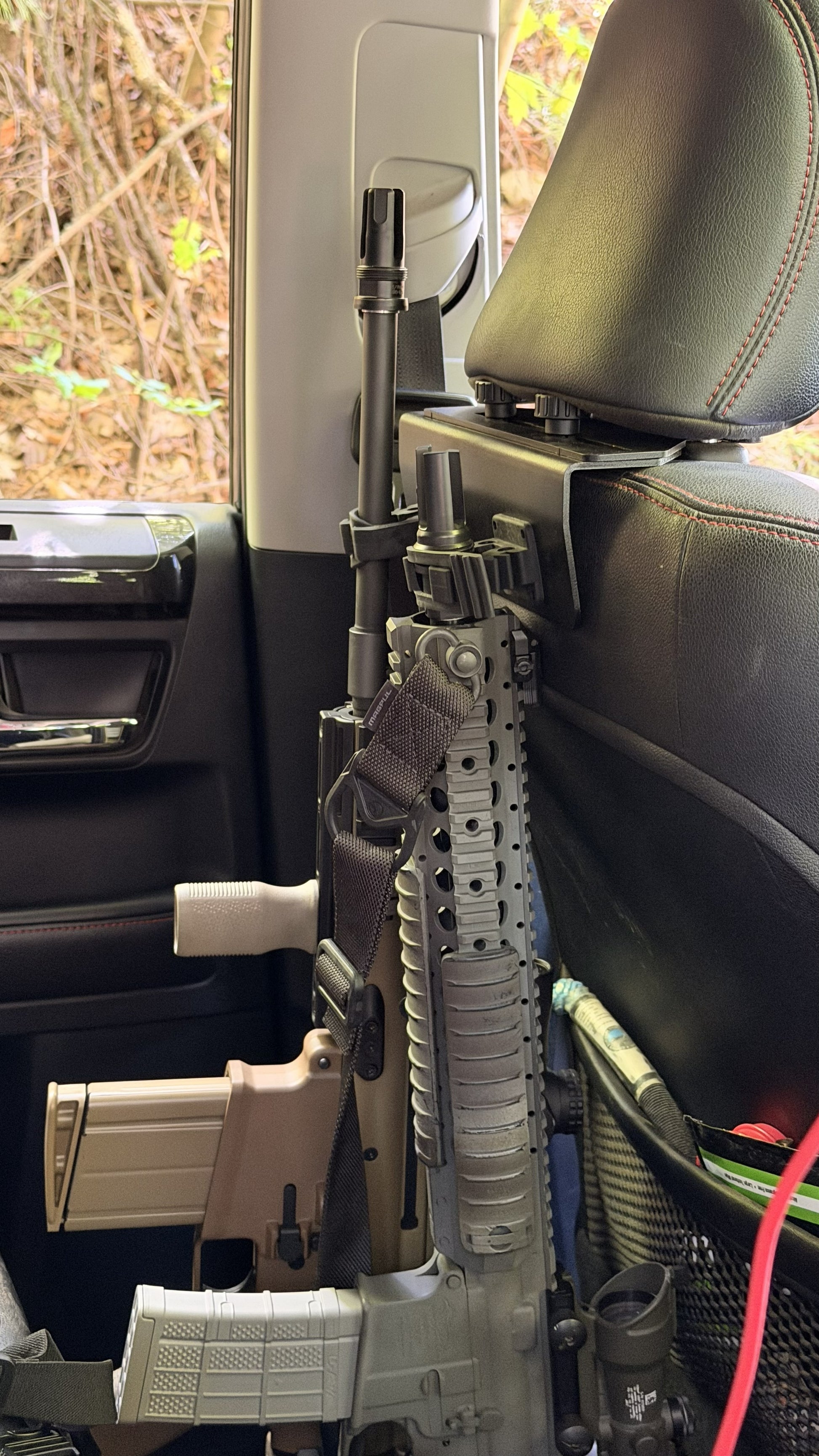 Two rifles are secured side by side to the back of a car’s front passenger seat using the Headrest Rifle Mount - HRM, with barrels pointing upward. The car’s interior, window, and shrubs outside are visible.