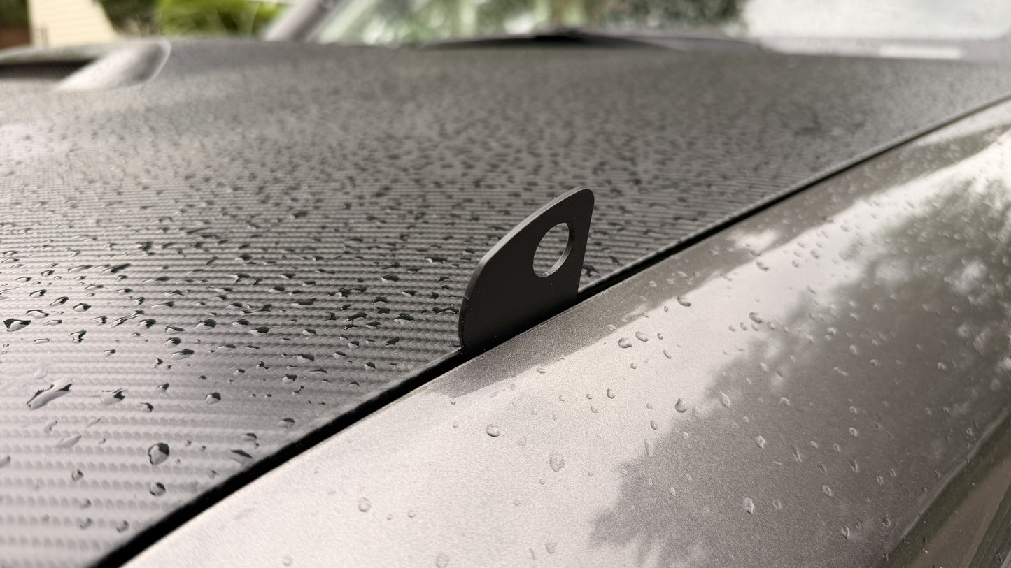 A close-up of Limb Riser Hood Brackets - 5th Gen 4Runner shows water droplets and a carbon fiber-like textured surface. A small metal tab with a hole protrudes from the gap between the hood and fender.