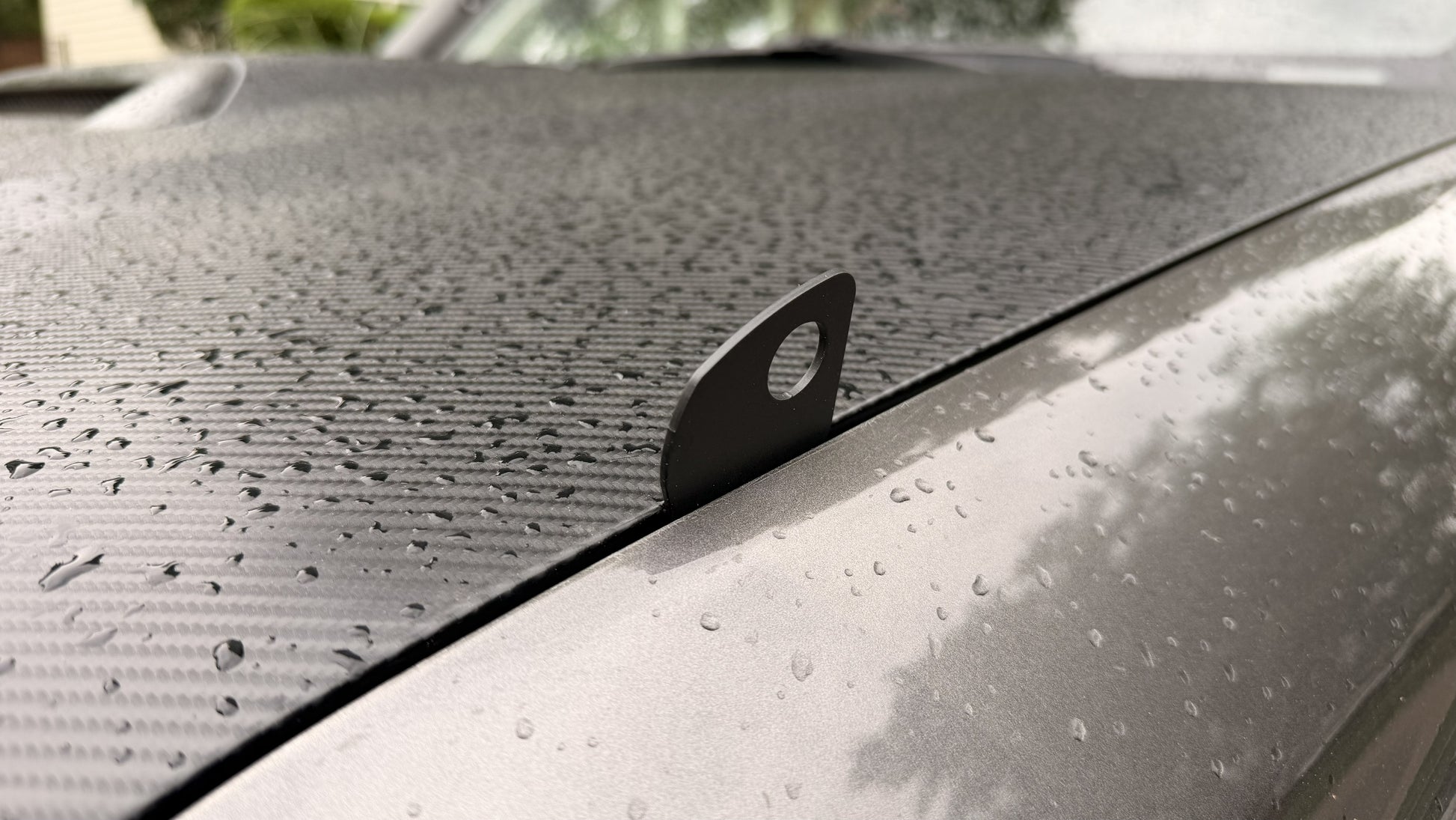 A close-up of Limb Riser Hood Brackets - 5th Gen 4Runner shows water droplets and a carbon fiber-like textured surface. A small metal tab with a hole protrudes from the gap between the hood and fender.