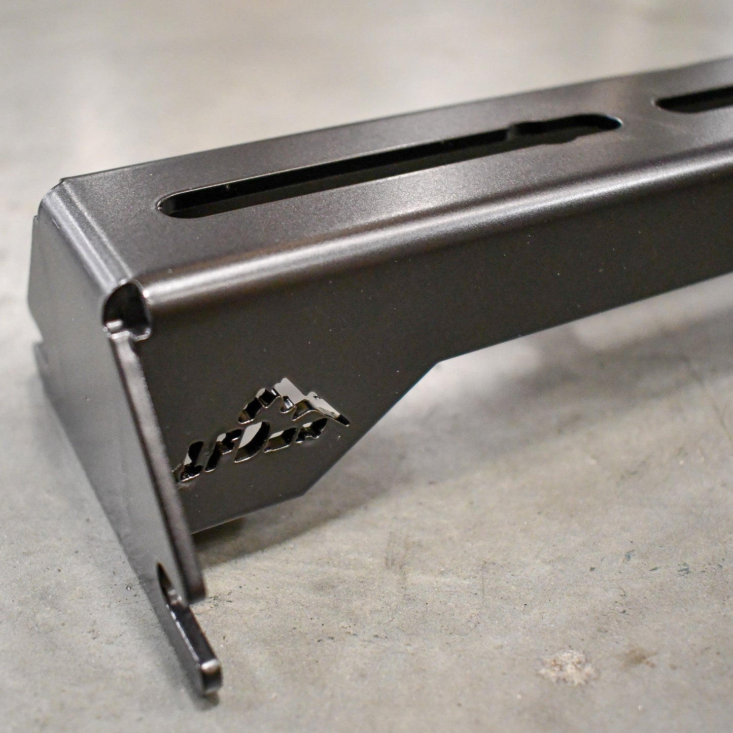 Close-up of a black metal bracket with slotted holes and a mountain logo from the Ruggedized Crossbar Kit - 5th Gen 4Runner, shown on concrete for factory roof rail storage solutions.