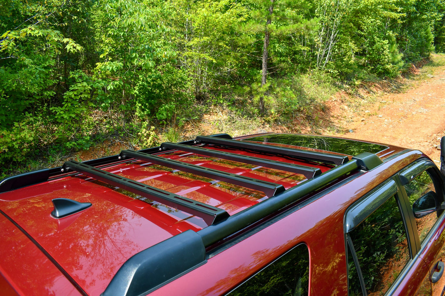 A close-up of the Ruggedized Crossbar Kit - 5th Gen 4Runner on factory roof rails of a red SUV, parked on a dirt road with lush greenery—a perfect roof storage solution for your next outdoor adventure.