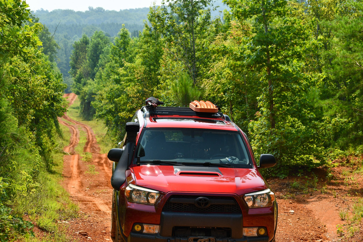 A red 5th Gen 4Runner equipped with the Ruggedized Crossbar Kit drives on a narrow dirt road through dense green forest under the sun, its off-road gear securely mounted for the adventure ahead.