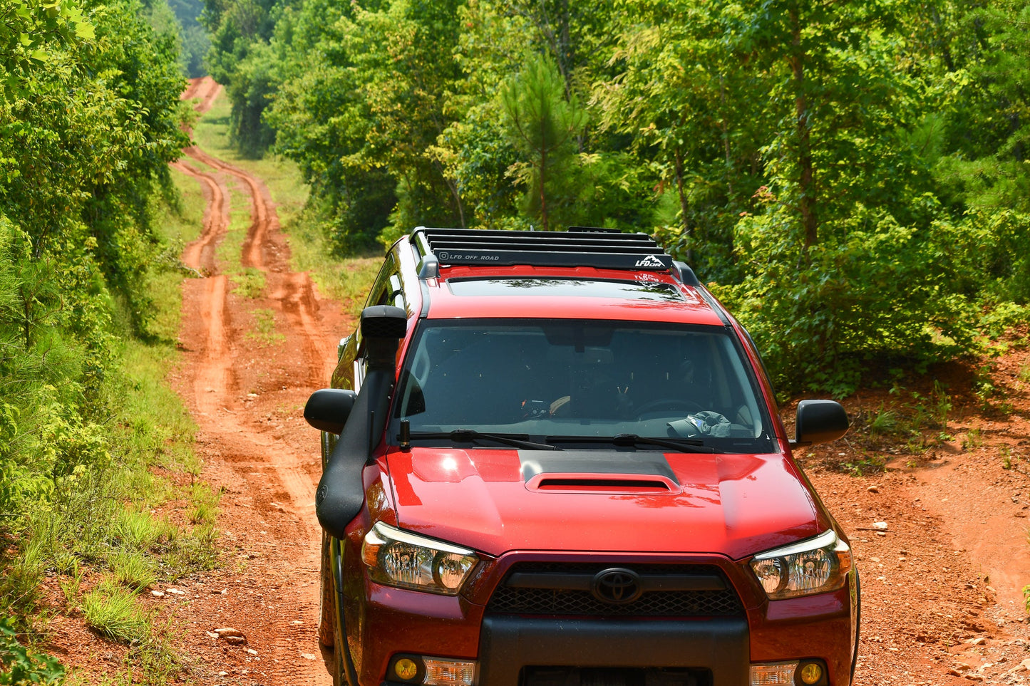A red Toyota SUV with a Ruggedized Crossbar Kit - 5th Gen 4Runner drives along a winding dirt road through a lush green forest, offering the ideal roof storage solution for adventure on sunny days.