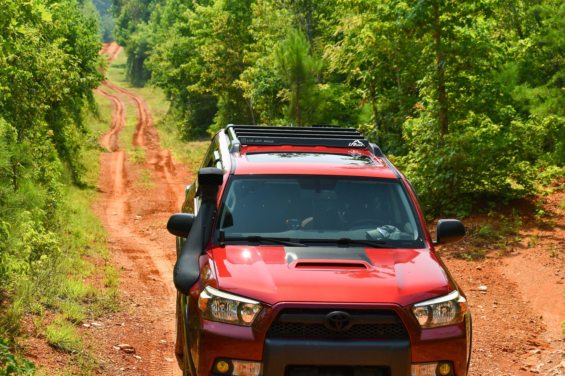 A red Toyota SUV with a Ruggedized Crossbar Kit - 5th Gen 4Runner drives along a winding dirt road through a lush green forest, offering the ideal roof storage solution for adventure on sunny days.