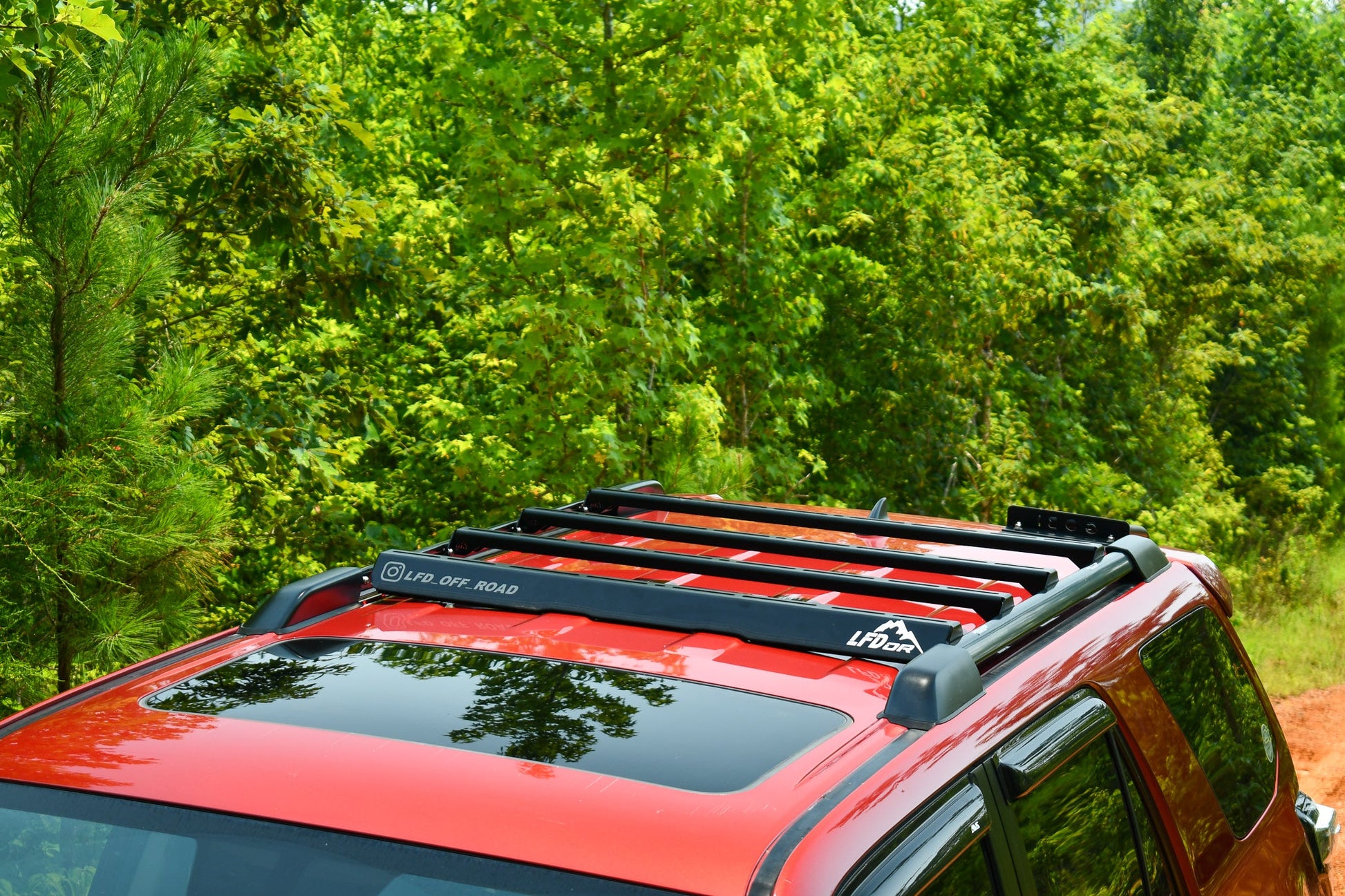 A red 5th Gen 4Runner with the Ruggedized Crossbar Kit is parked on a dirt path amid lush trees, delivering dependable roof storage for any sunny day adventure.