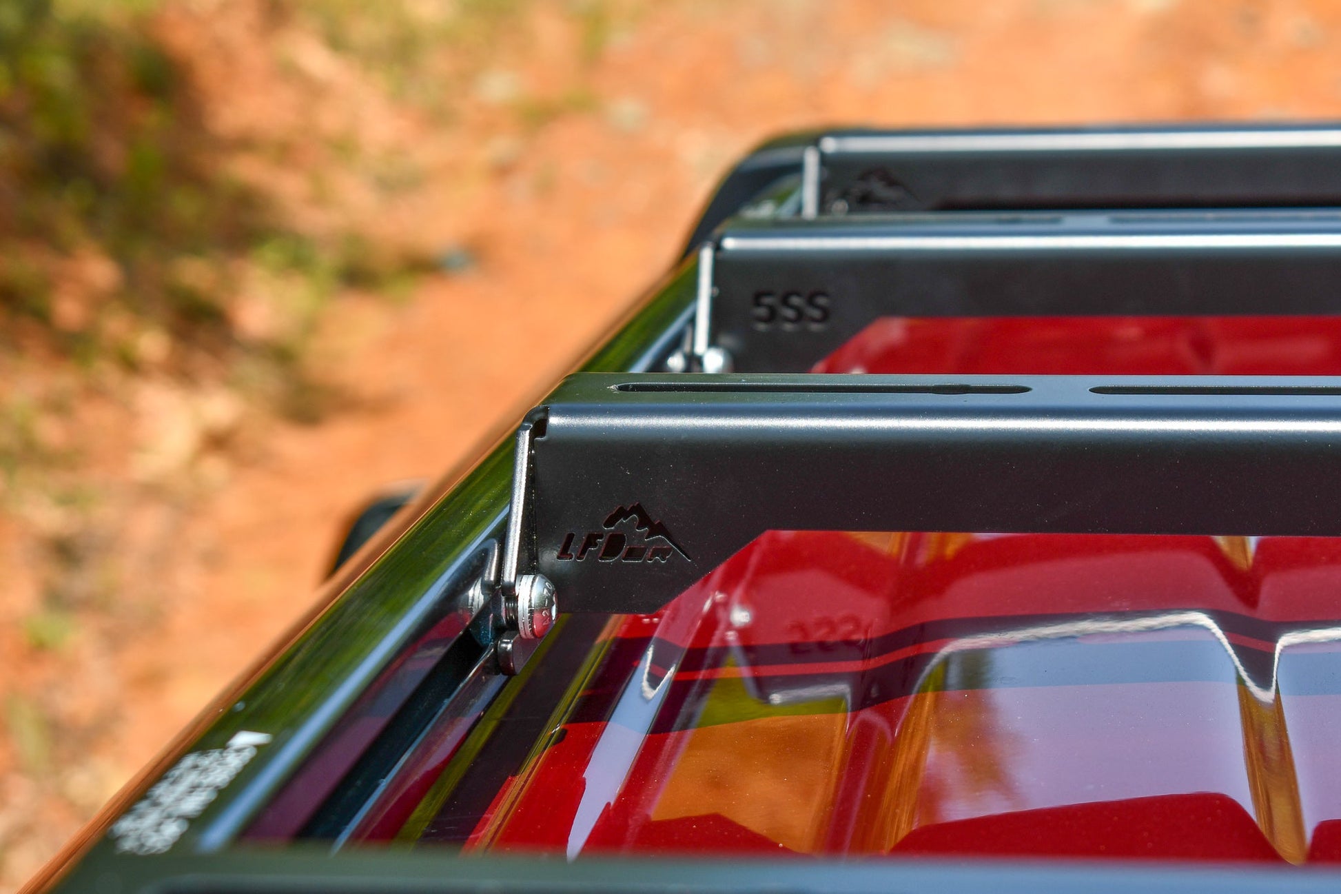 Close-up of a black metal Ruggedized Crossbar Kit - 5th Gen 4Runner mounted on a red vehicle, bolts and LFD logo visible. Blurred outdoor backdrop with dirt path and greenery—ideal roof storage for your 4Runner adventures.